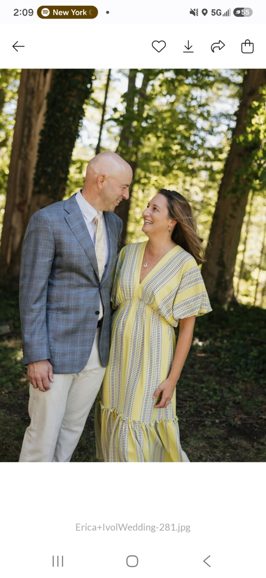 A man and woman stand outdoors in a wooded area, smiling at each other, with trees and sunlight in the background.