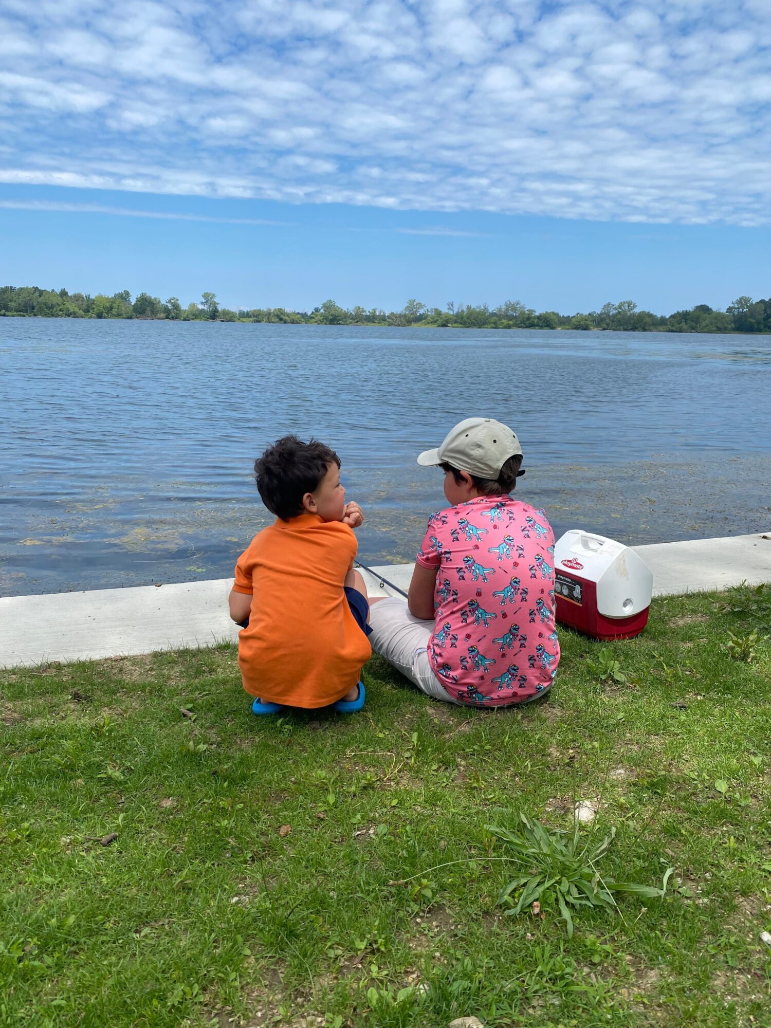 Two people sit on the grass near a body of water, with a cooler beside them, under a partly cloudy sky.
