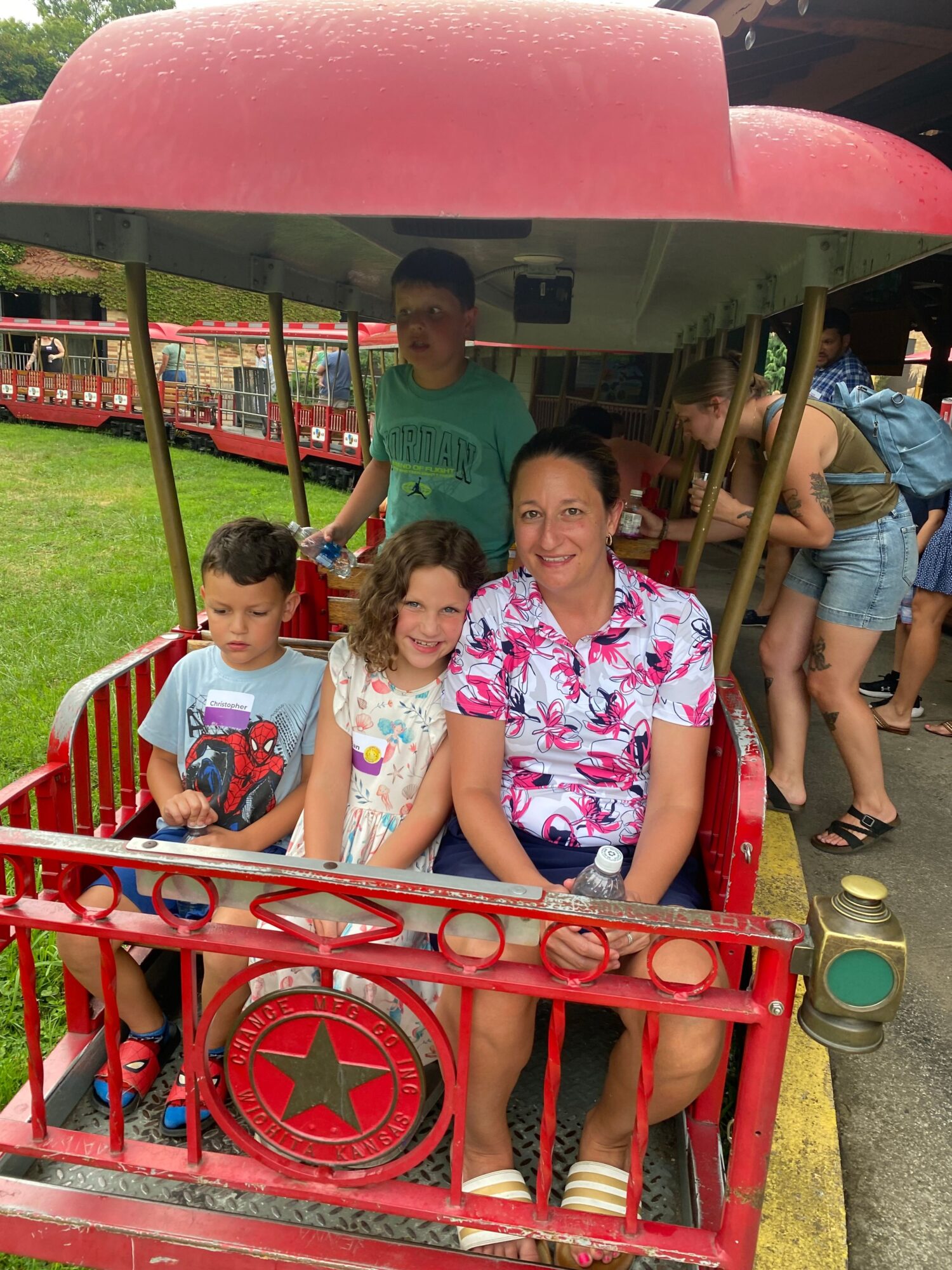 Group of children sitting on a red amusement ride, with some standing and others seated, outdoors on a sunny day.