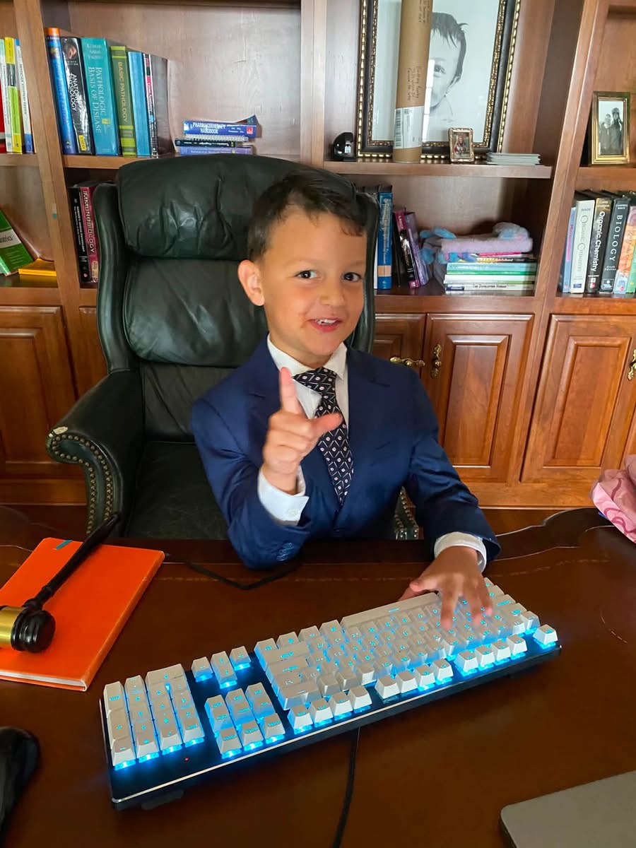 Young boy in a suit sitting at a desk, pointing, with a keyboard and bookshelves in the background.