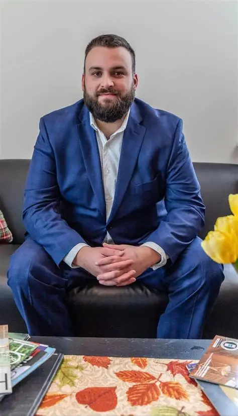 Man in blue suit sitting on a black couch with hands clasped, in front of a table with a floral tablecloth and yellow flowers.
