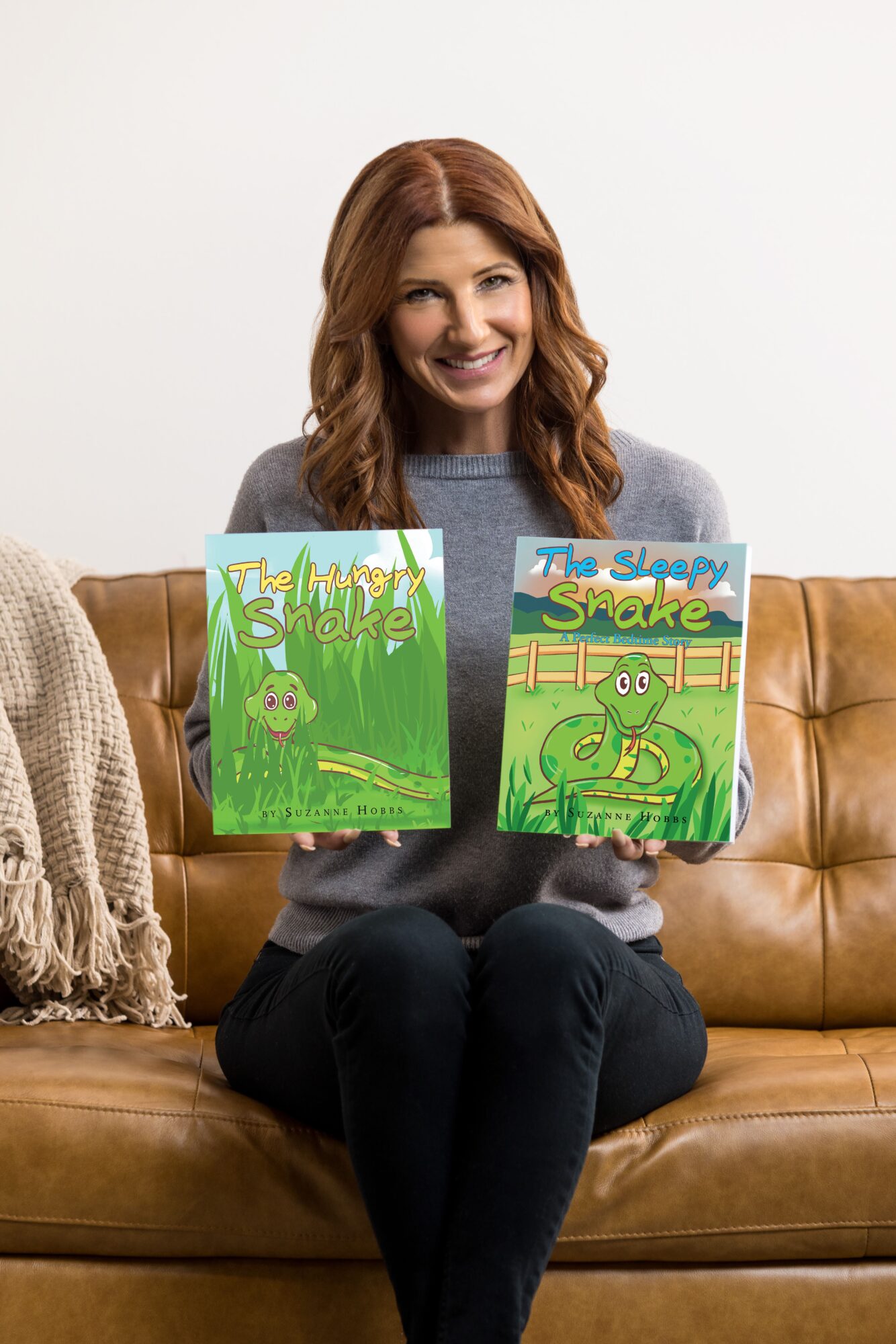 Woman with red hair holding two children's books, sitting on a brown couch, smiling, against a plain white wall.