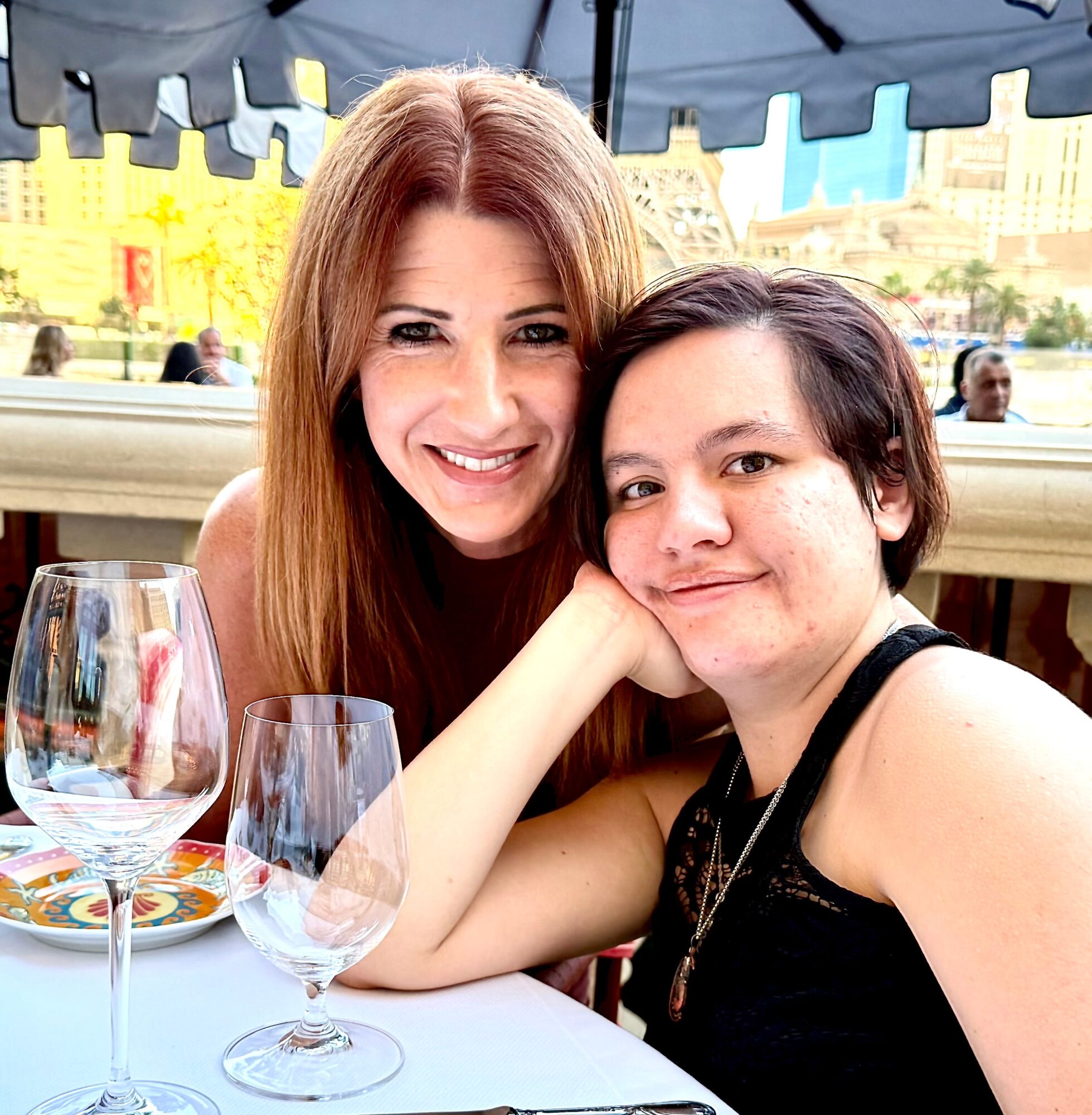Two women smiling at a table outdoors, with wine glasses and a cityscape background.