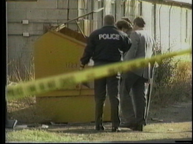 Police officer and two people near a large yellow dumpster, with yellow caution tape in the foreground.