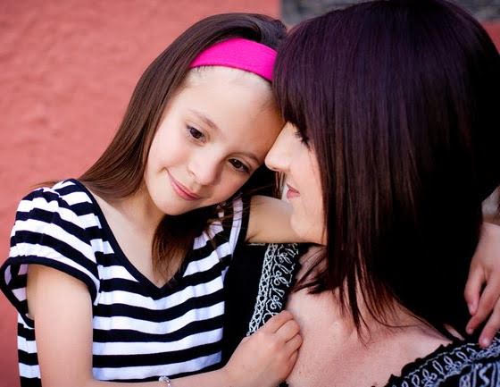Young girl with a pink headband leaning her head on an adult woman's shoulder, both smiling gently, close-up shot.