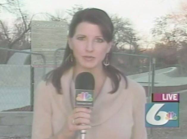 Female news reporter holding a microphone outdoors, live broadcast, trees and a fence in the background.
