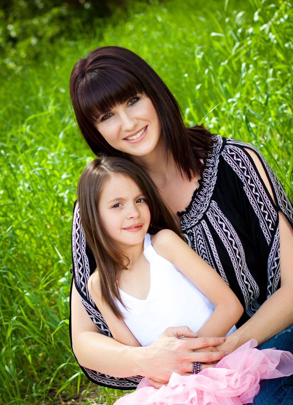 A woman and a girl smiling outdoors with green foliage in the background.