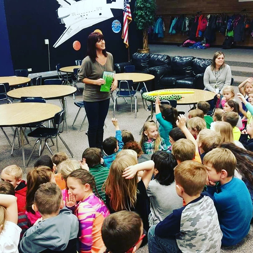 Woman standing in front of children in a classroom, holding a green book, with children raising hands and sitting on the floor.