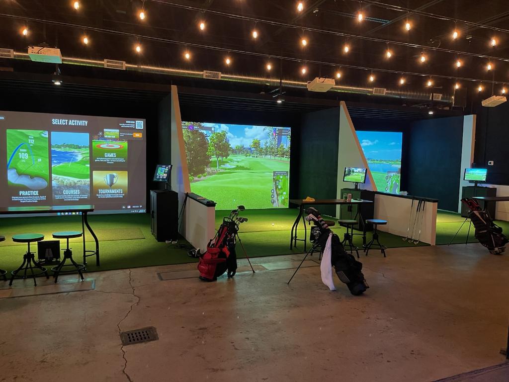 Indoor golf simulation room with three screens displaying virtual golf courses, golf bags, and equipment, illuminated by string lights.