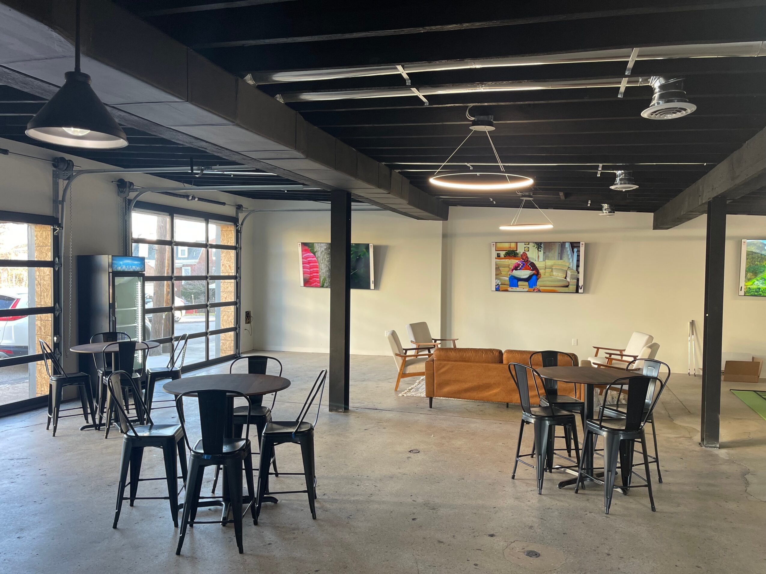 Interior of a cafe with tables, chairs, large windows, and wall art, under a black ceiling with hanging lights.