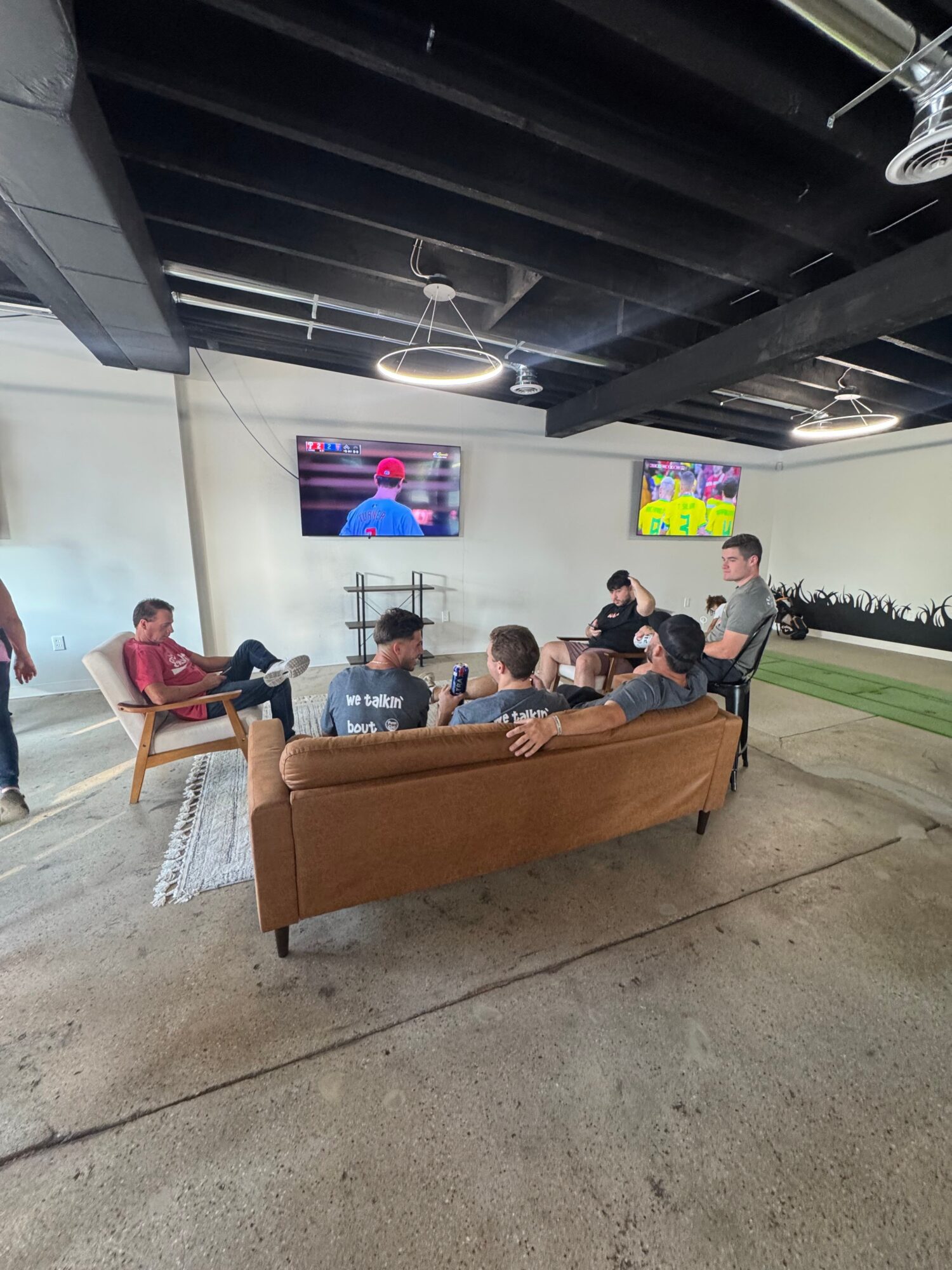 Group of people sitting on a couch in a waiting area, with a TV on the wall and a colorful poster, seen from the side.