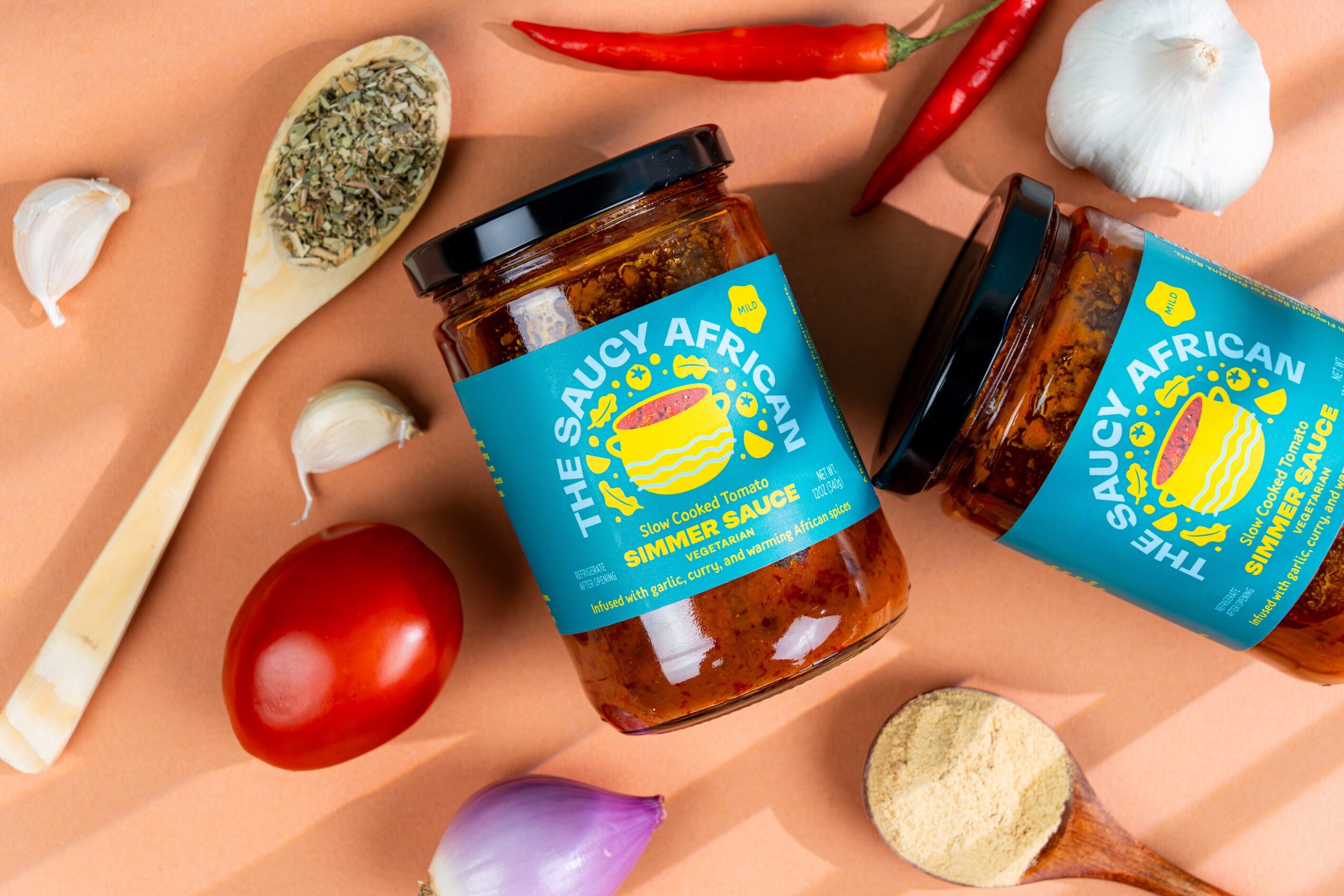 Two jars of African sauce surrounded by garlic, chili peppers, a tomato, and a wooden spoon with seasoning, on a peach background.