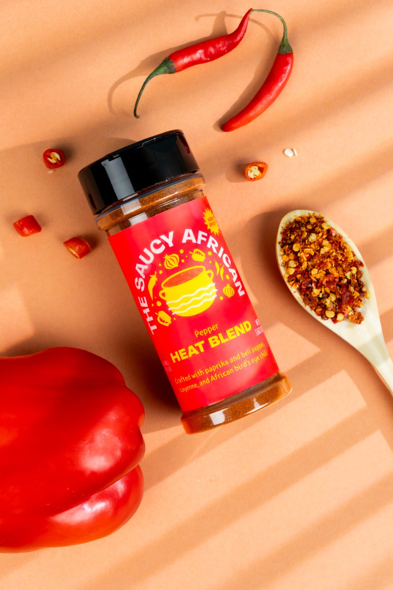 Bottle of spicy heat blend seasoning surrounded by red chili peppers, a red bell pepper, and a spoonful of seasoning on a peach background.