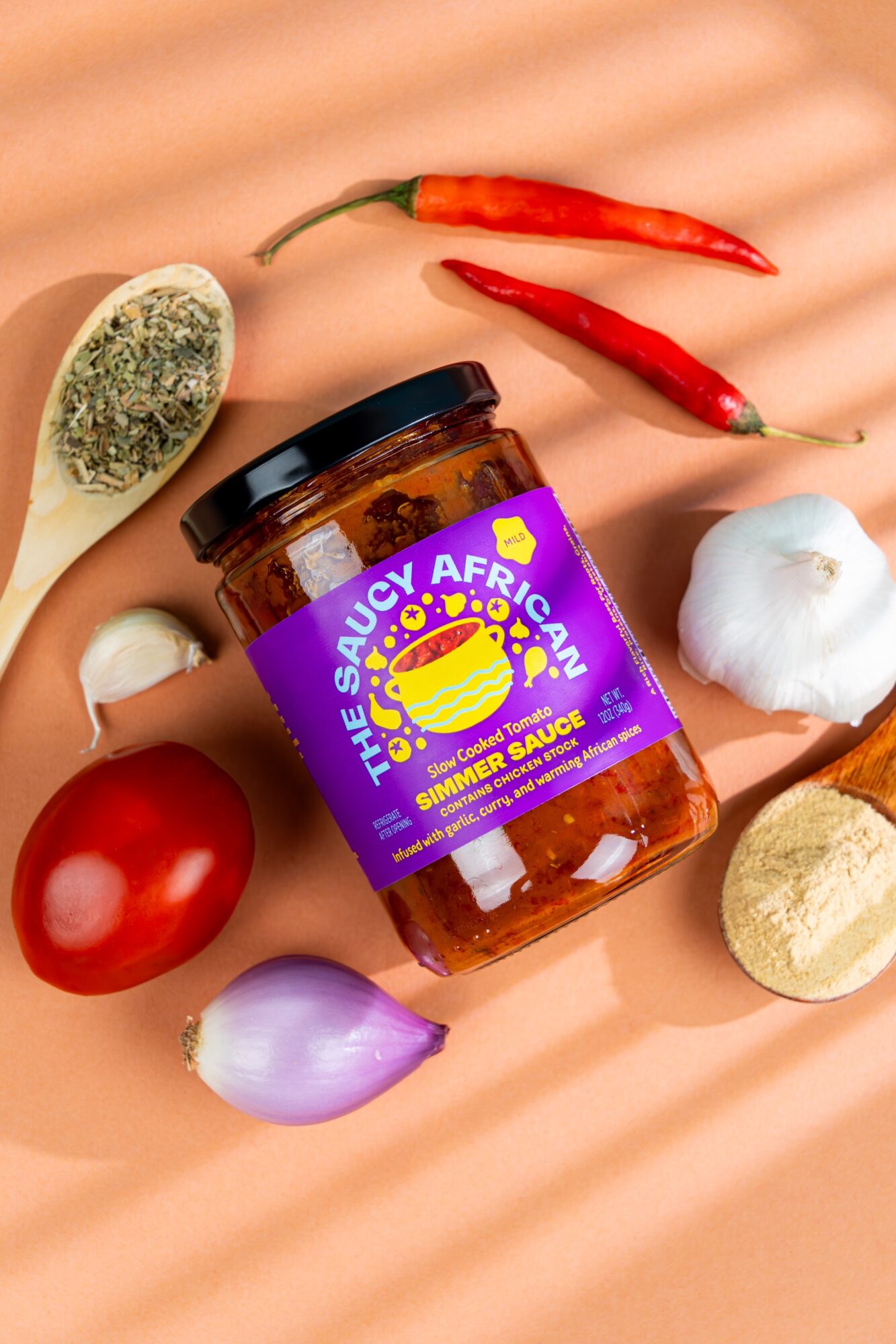 Jar of African sauce surrounded by chili peppers, garlic, onion, tomato, and shallots on a peach-colored surface.