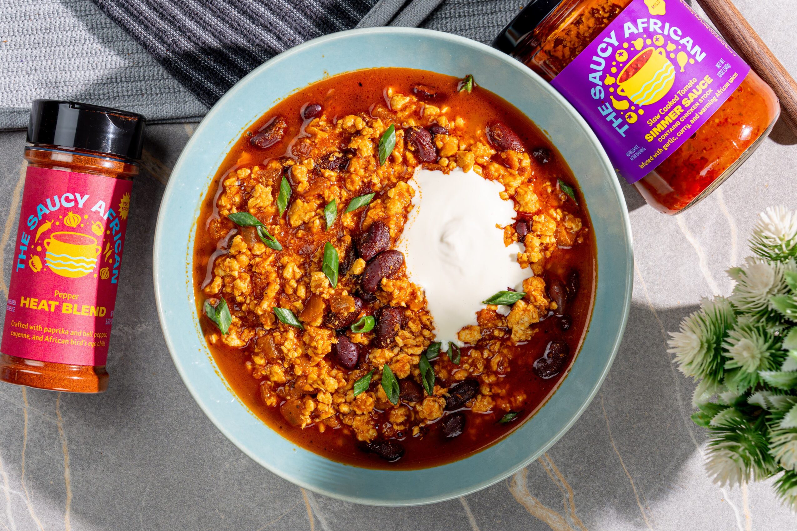Bowl of chili with beans, topped with sour cream, surrounded by spice jars and a plant, on a gray surface.