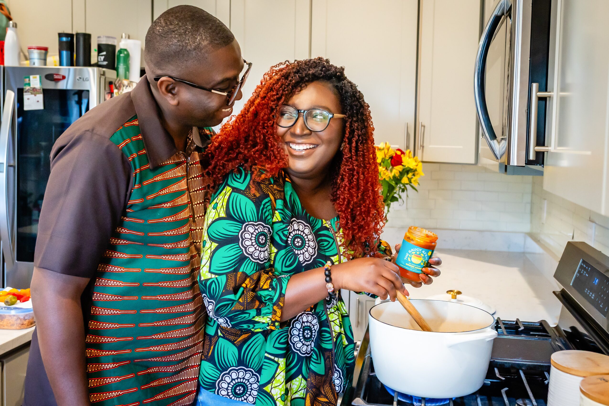 Two women in a kitchen, one holding a jar, smiling at each other, with a pot on the stove and flowers in the background.