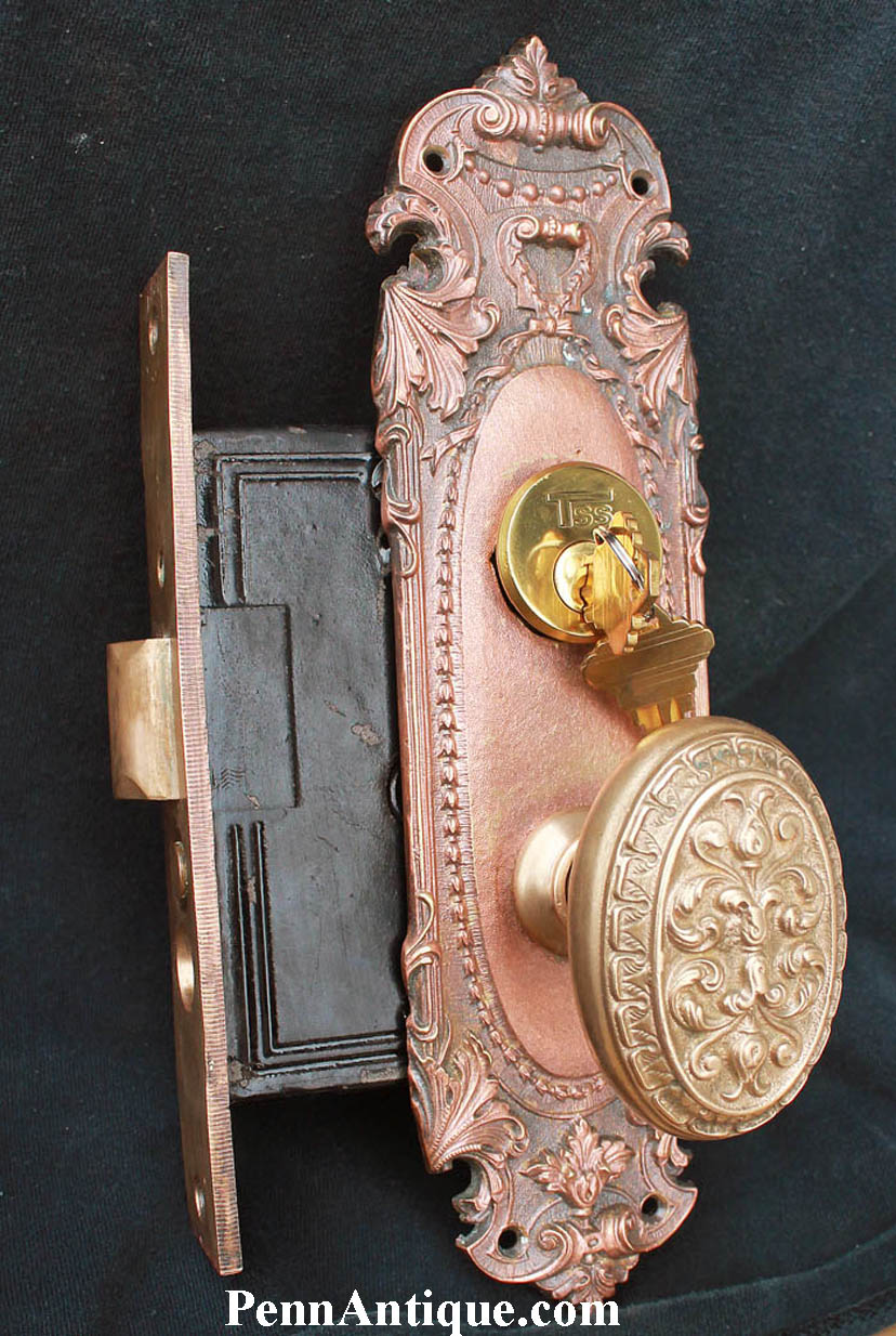 Decorative antique door lock with ornate metalwork and a round handle, mounted on a dark background.