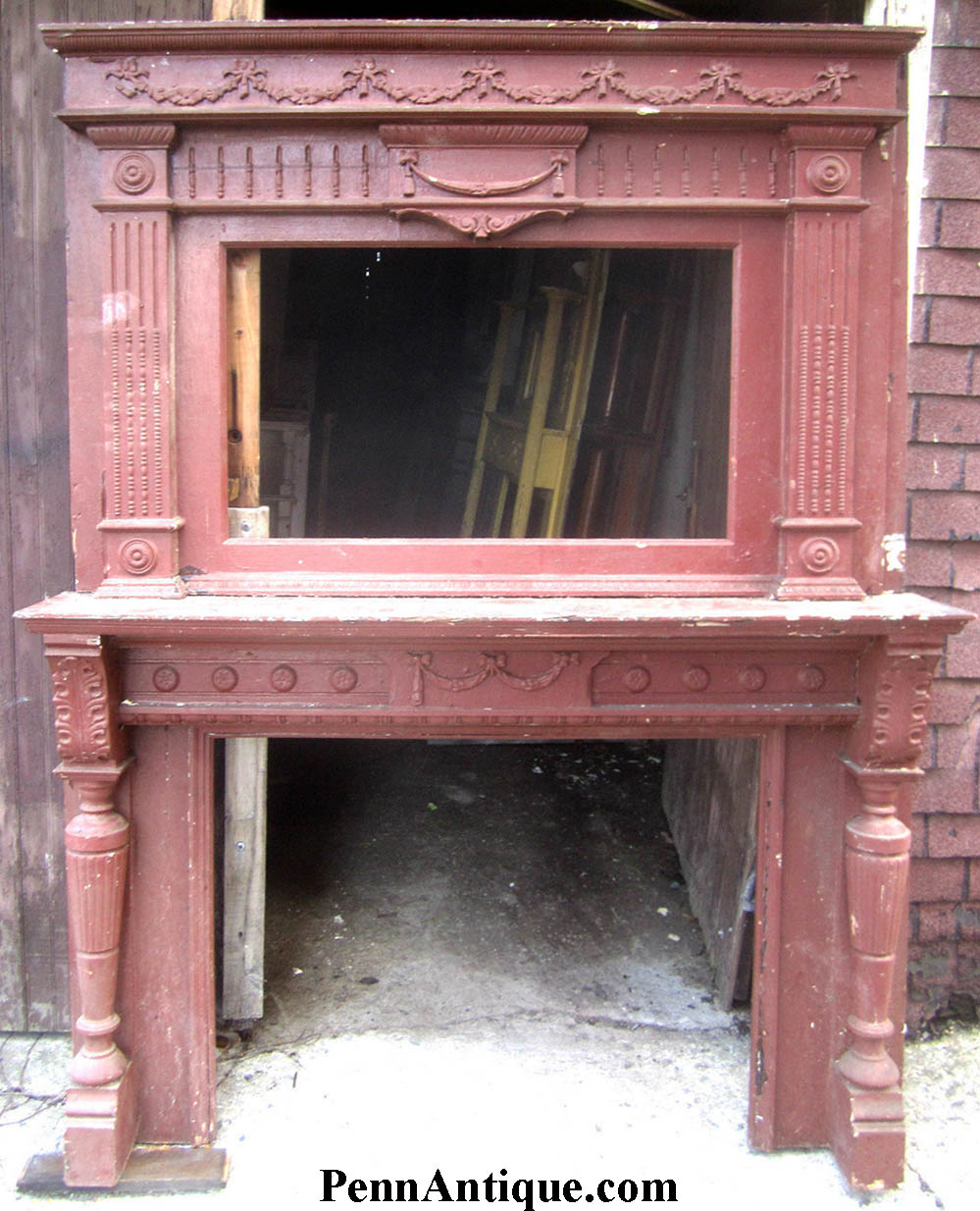 Decorative wooden fireplace mantel with mirror, detailed carvings, and columns, placed outdoors against a brick wall.