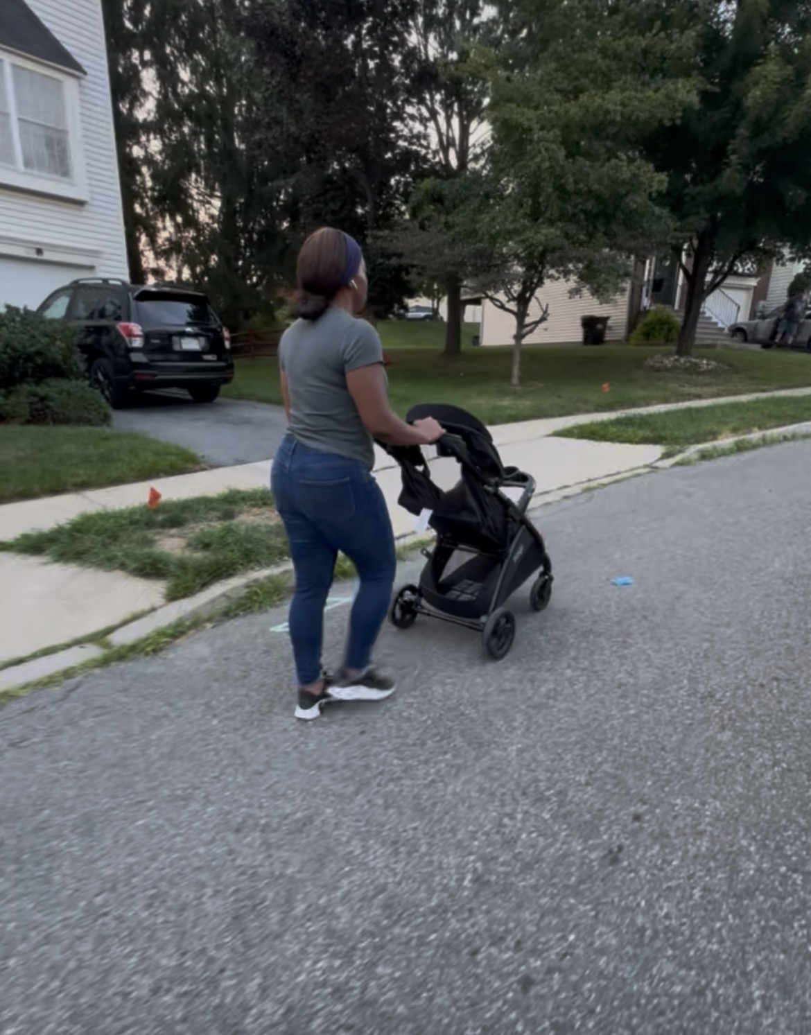 Woman with short hair pushing a stroller on a suburban street, trees and houses in background.