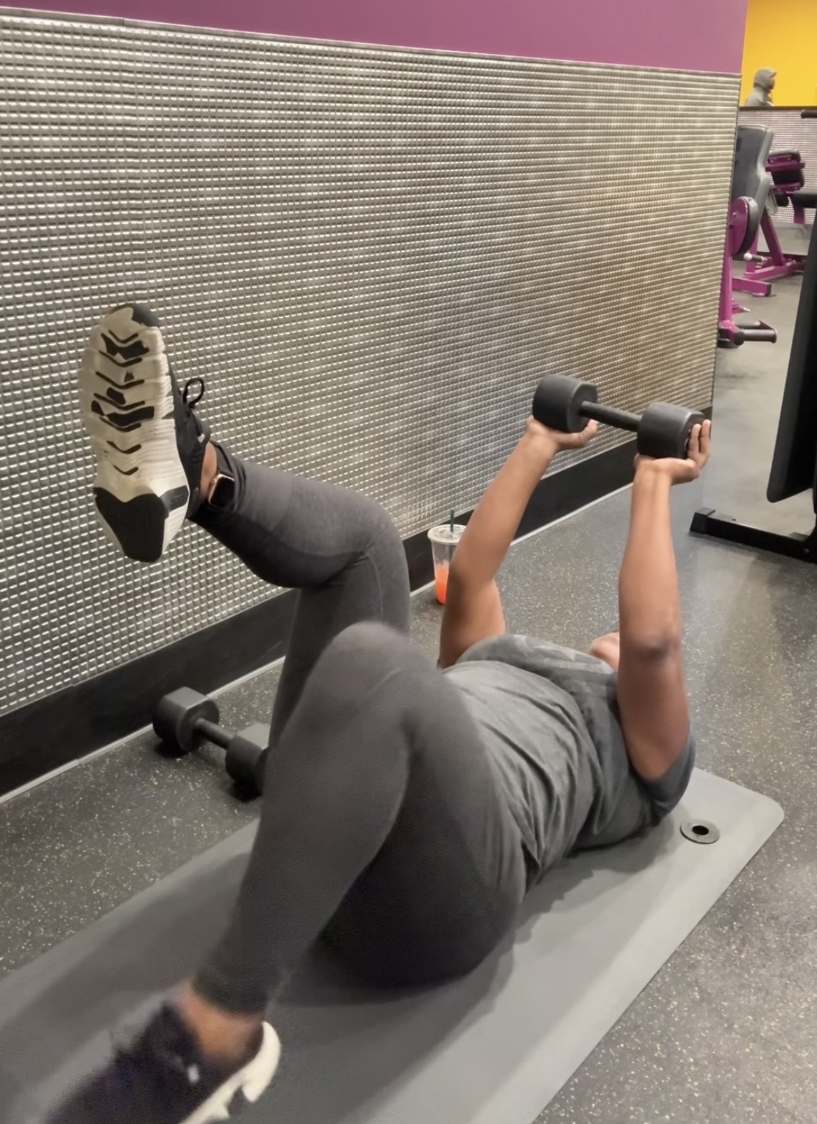 Person lying on a mat in gym, lifting dumbbell with one arm, wearing gray workout clothes, with gym equipment in background.
