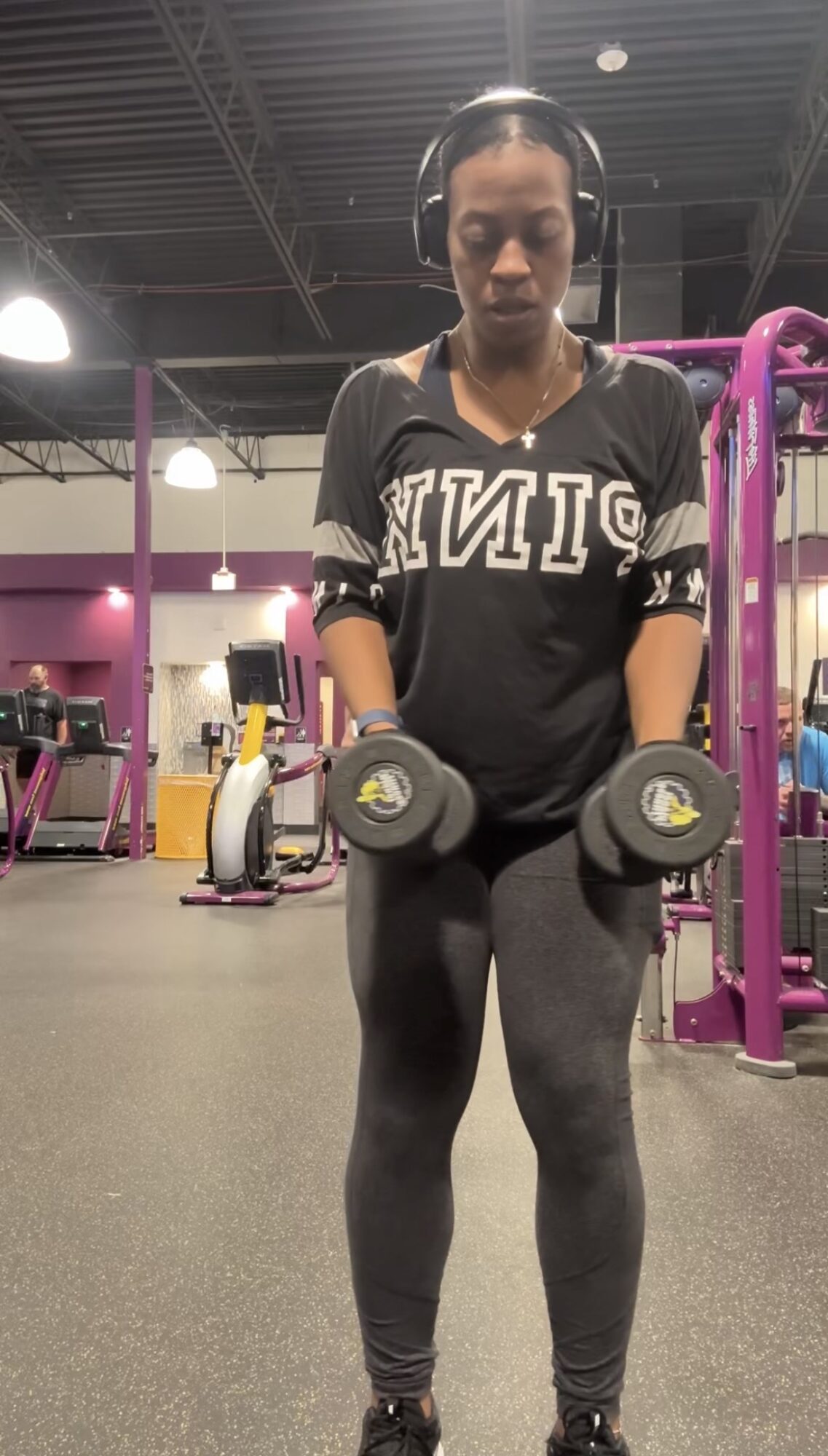 Woman in gym holding dumbbells, wearing headphones, black sweatshirt, and leggings, with gym equipment in background.