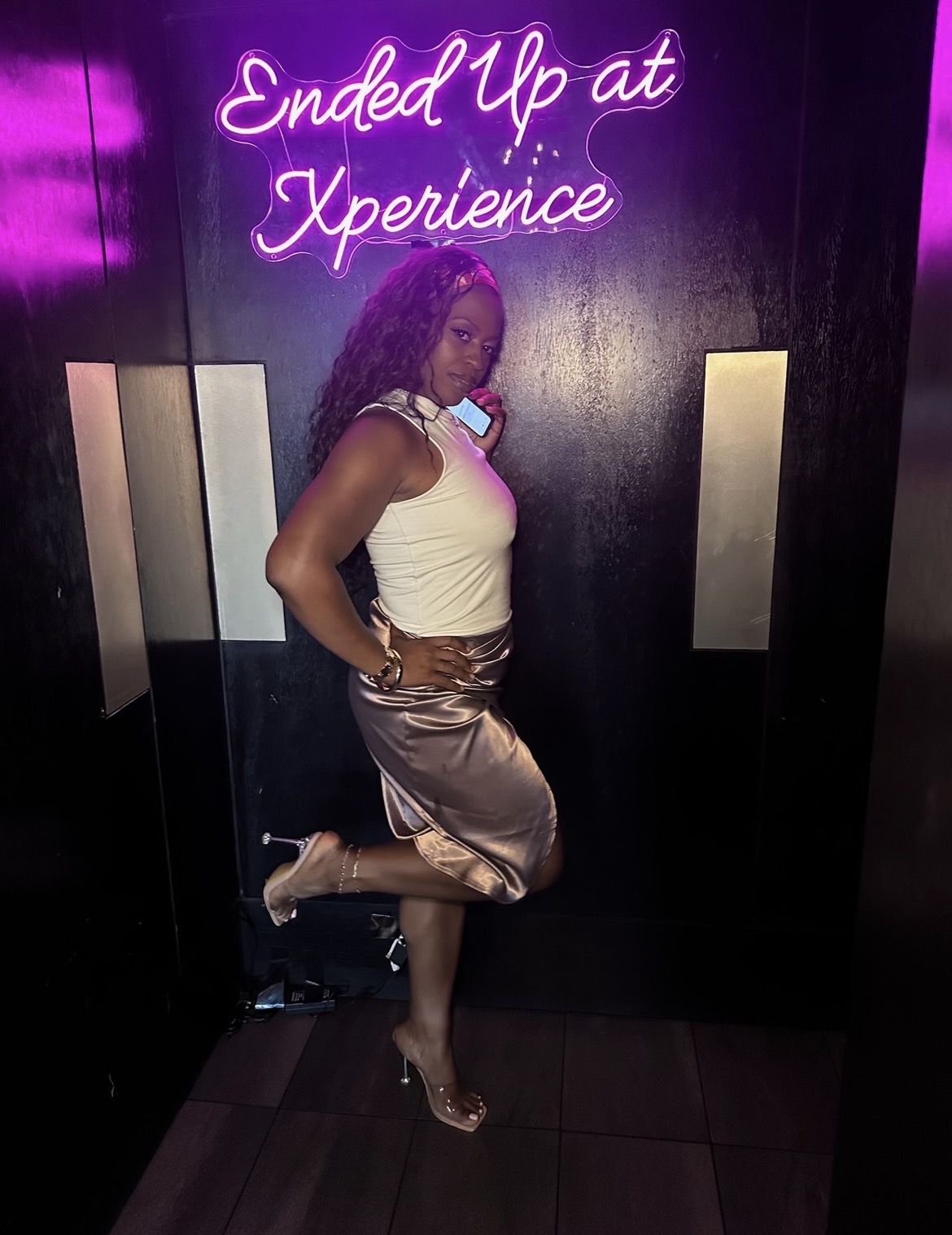 Woman with curly hair in a white top and satin skirt poses in front of a neon sign, standing on one leg.