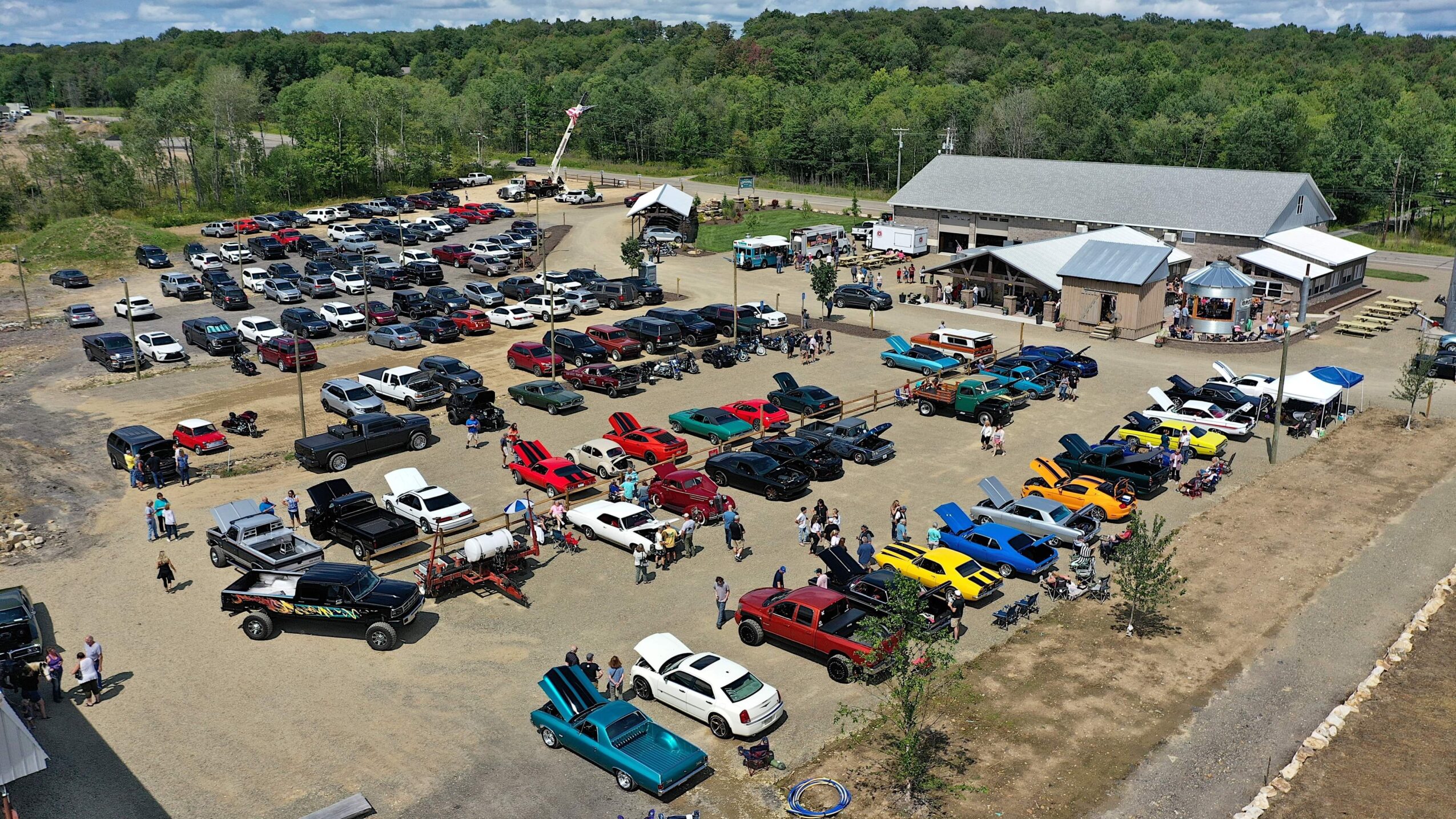 Aerial view of a large parking lot filled with classic cars and trucks, adjacent to a building with a crowd outside.