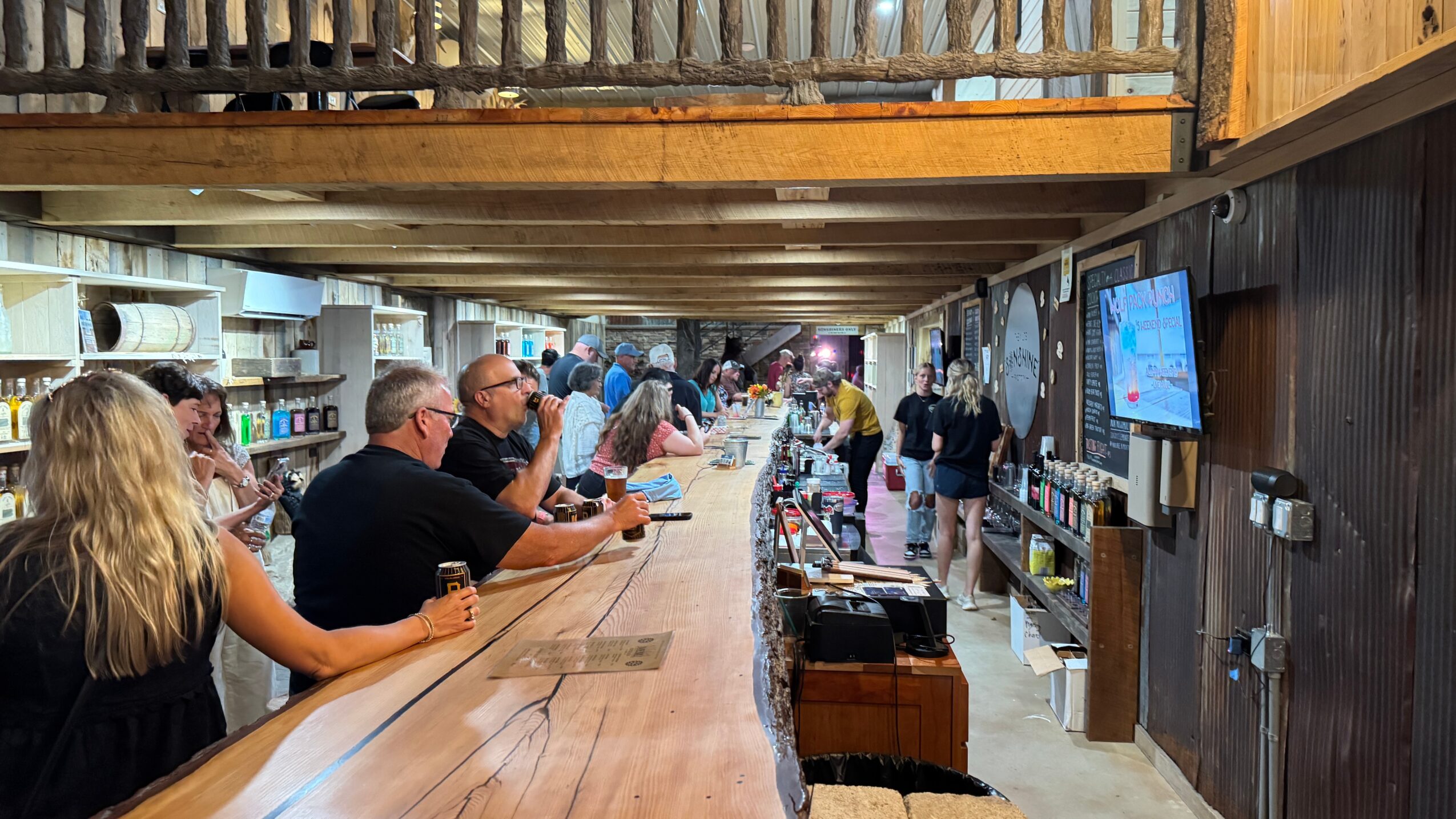 People sit at a long bar counter in a busy indoor space with shelves and a television screen on the wall.