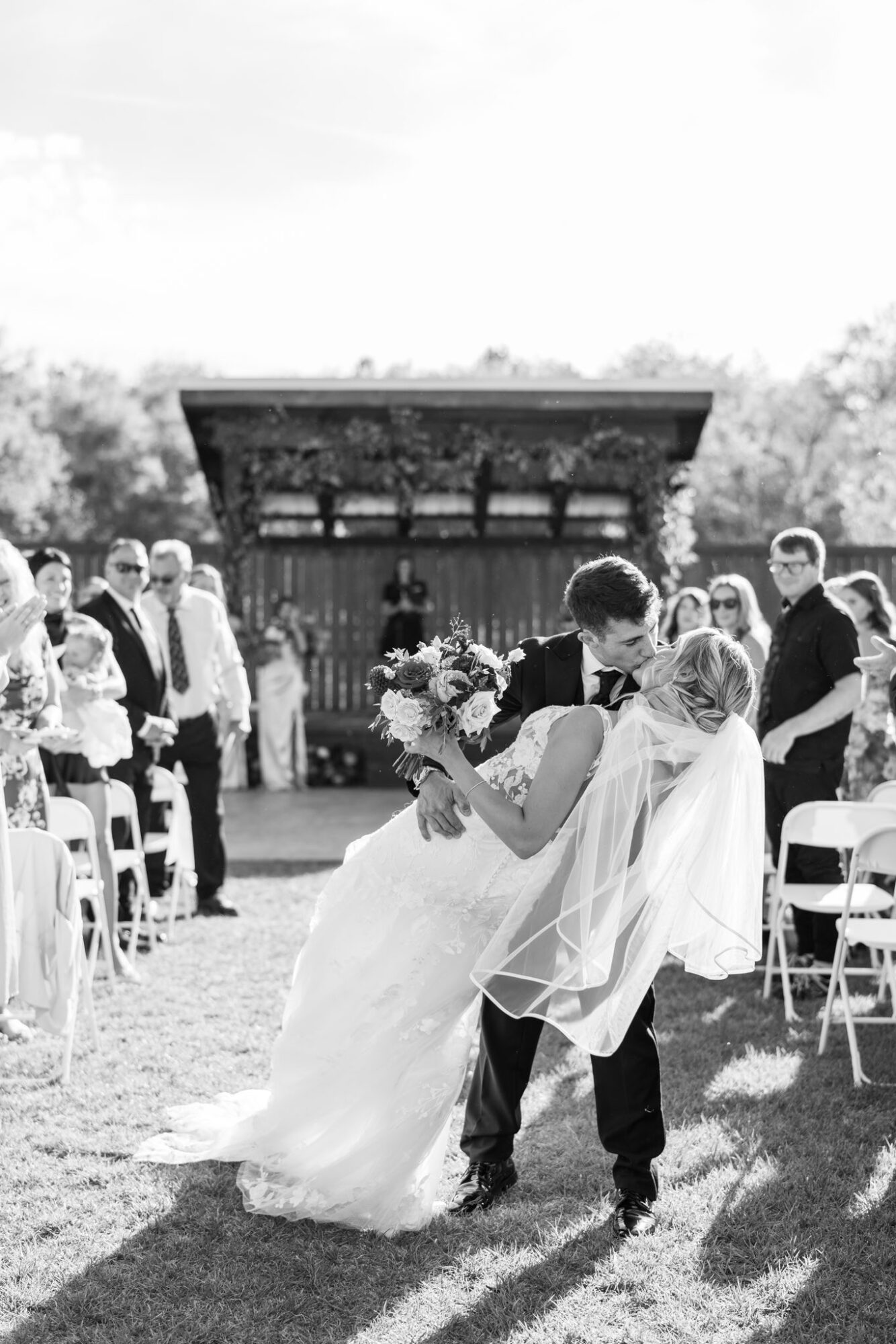 Couple dancing at outdoor wedding ceremony with guests and a pavilion in background, sunlight casting shadows.