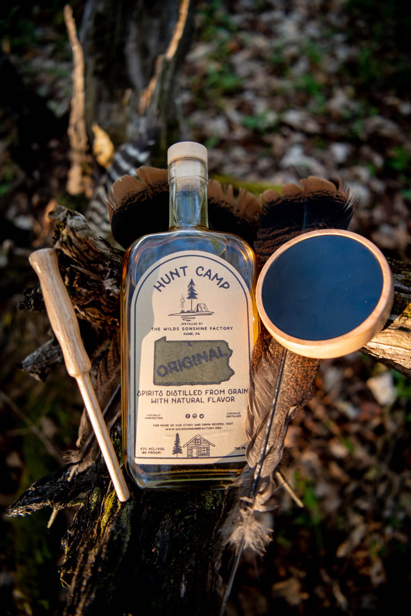 A bottle of Hunt Camp Original syrup with a spoon and a round container on a tree stump outdoors.