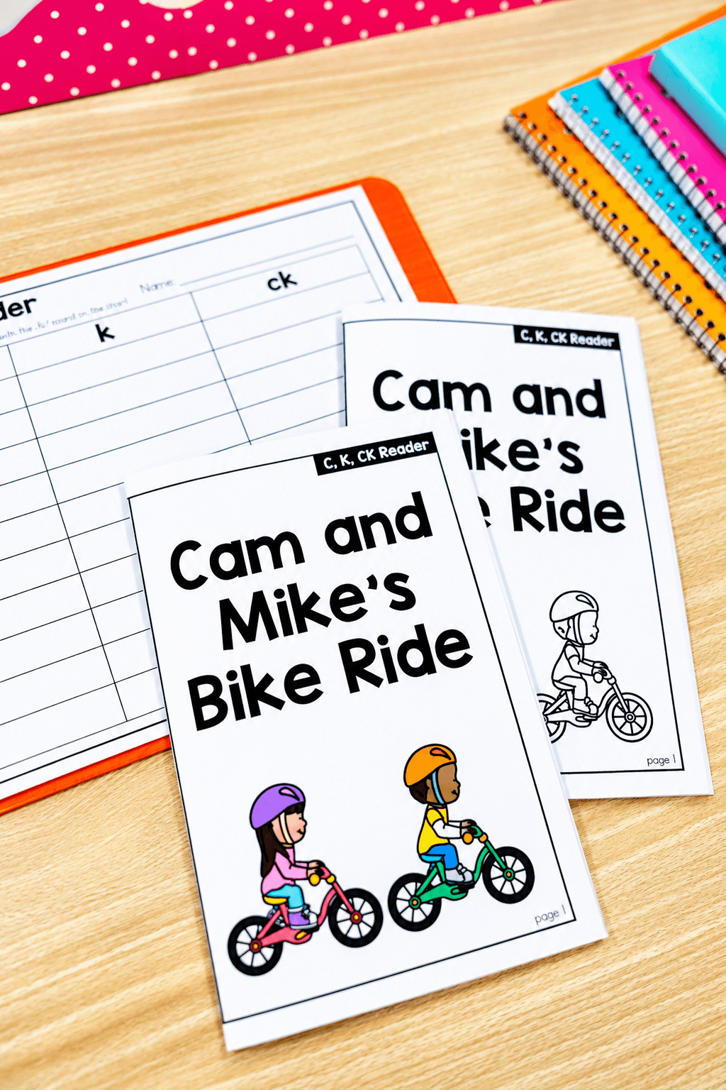 Two booklets titled 'Cam and Mike's Bike Ride' with illustrations of children riding bikes, on a wooden desk with colorful notebooks.