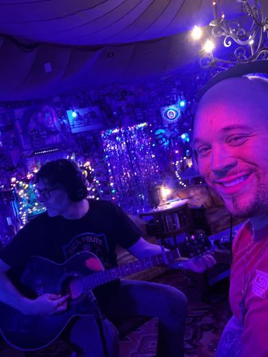 Two people in a dimly lit room with colorful lights, one playing guitar and the other smiling at the camera.