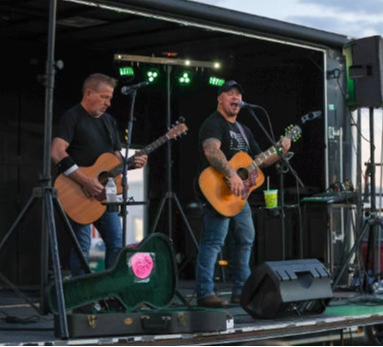 Two musicians playing guitars on an outdoor stage with microphones and equipment, under a partly cloudy sky.
