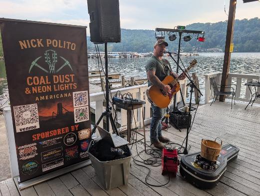 Musician playing guitar on a deck near a body of water with a sign advertising Nick Polito's music event.