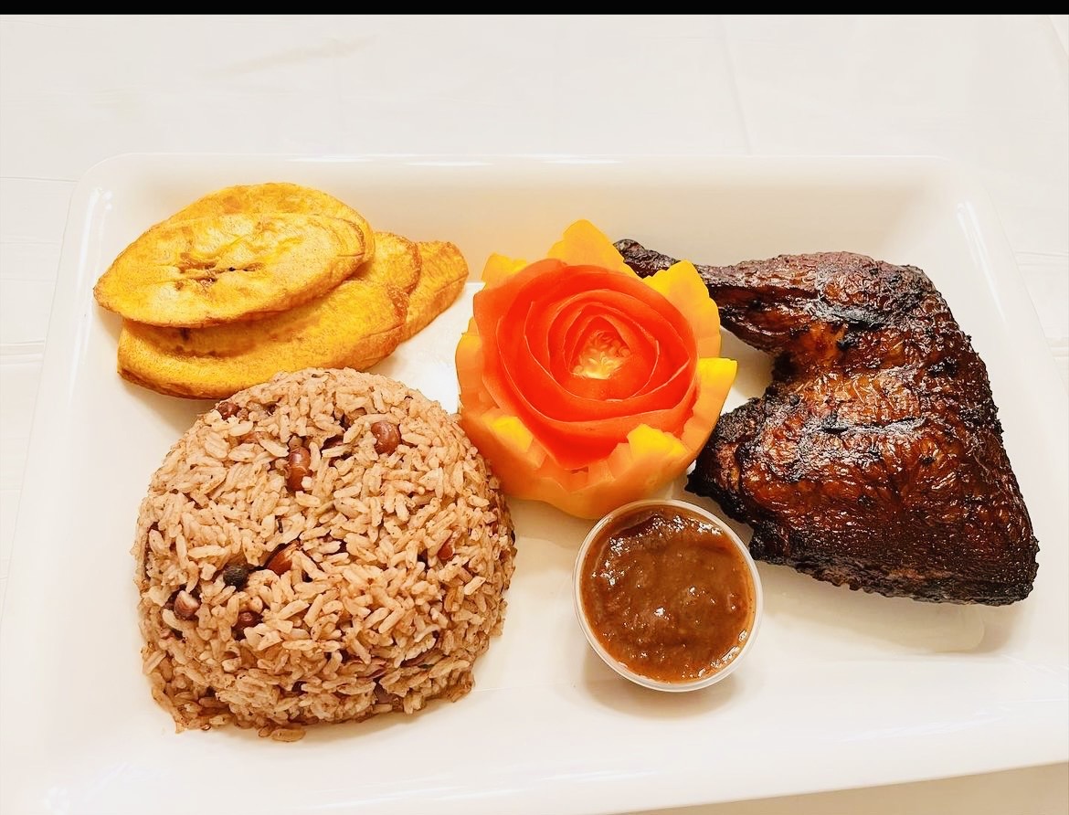 Plate with rice, grilled meat, fried plantains, and a carved vegetable garnish, with a small sauce container.
