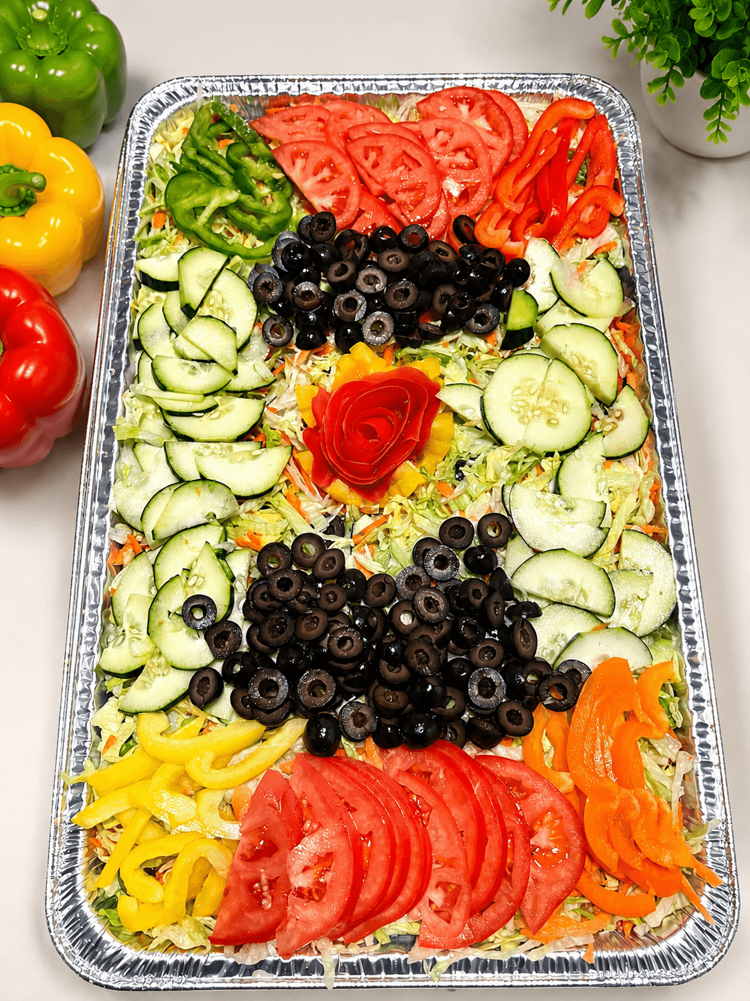 Tray of sliced cucumbers, tomatoes, black olives, yellow bell peppers, and carrots with a rose-shaped garnish, on a white surface.