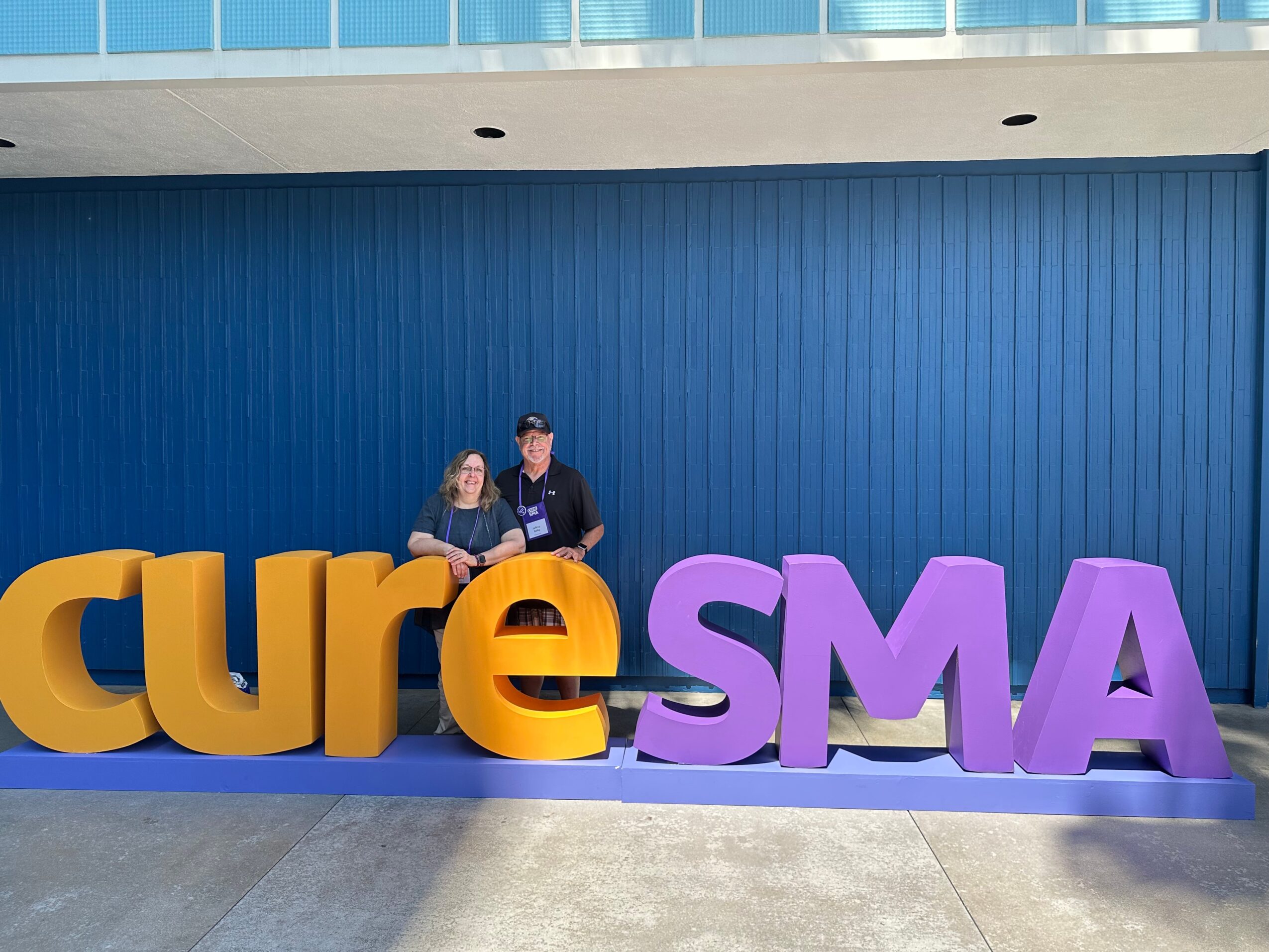 Two people sitting behind large colorful letters spelling 'cureSMAS' in front of a blue wall.