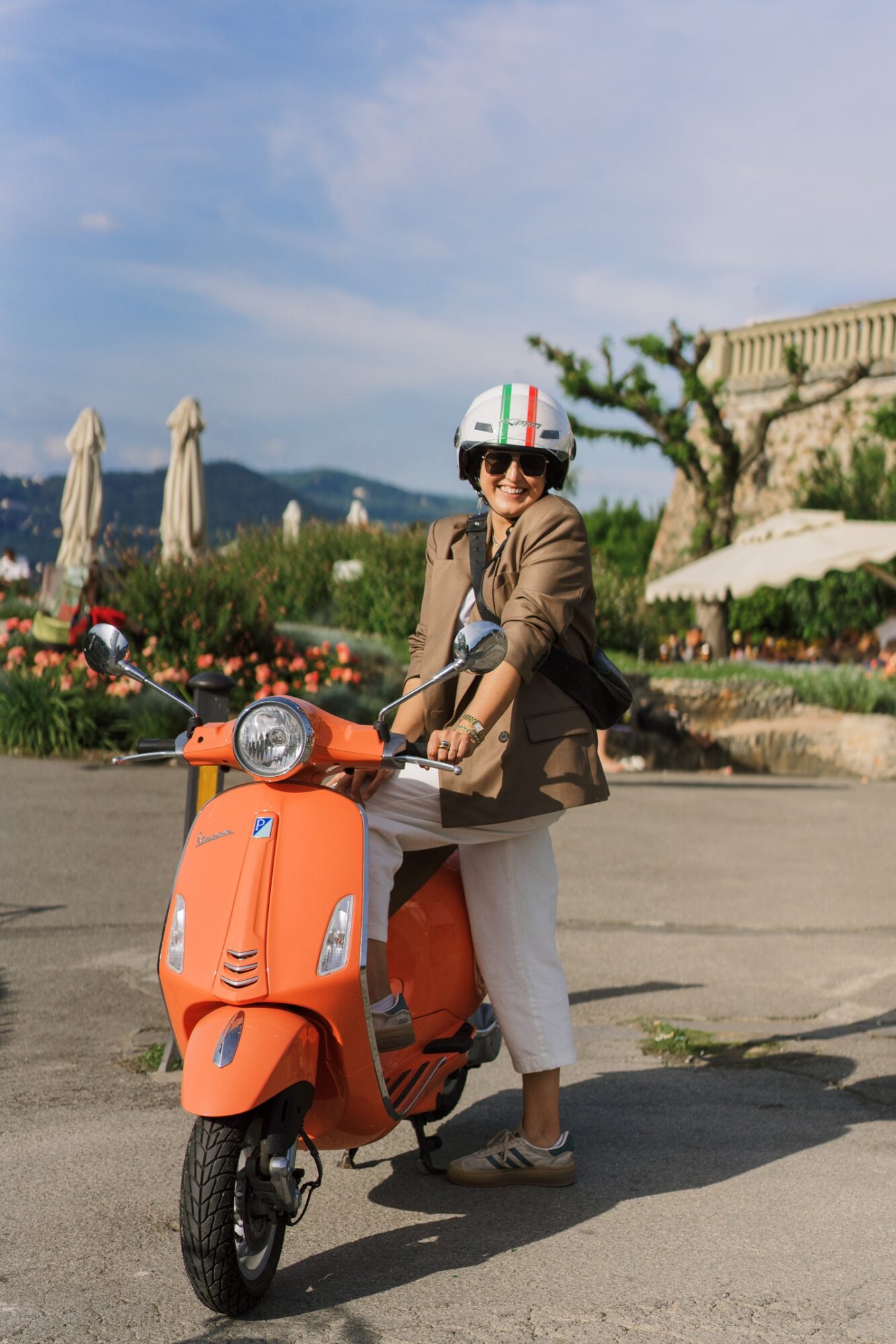 Person wearing sunglasses and a helmet, sitting on an orange scooter outdoors, with umbrellas and greenery in the background.