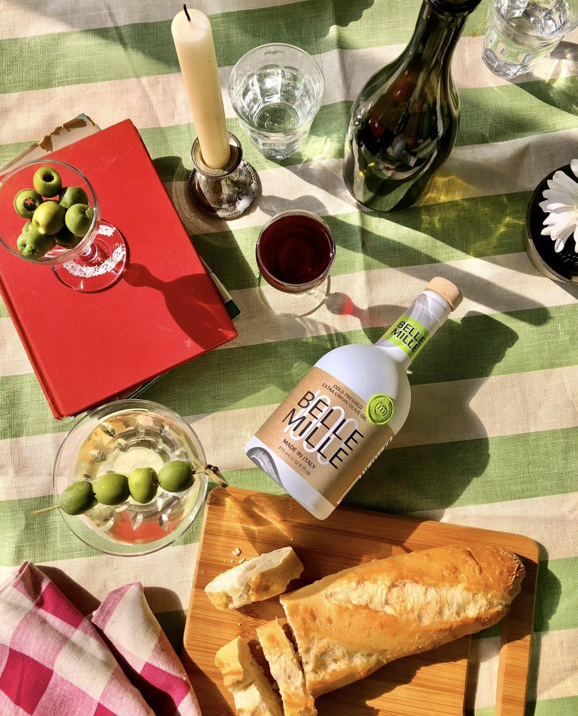 Table with bread, green olives, a bottle, a glass of red wine, a candle, and a red book on a green and white striped tablecloth.