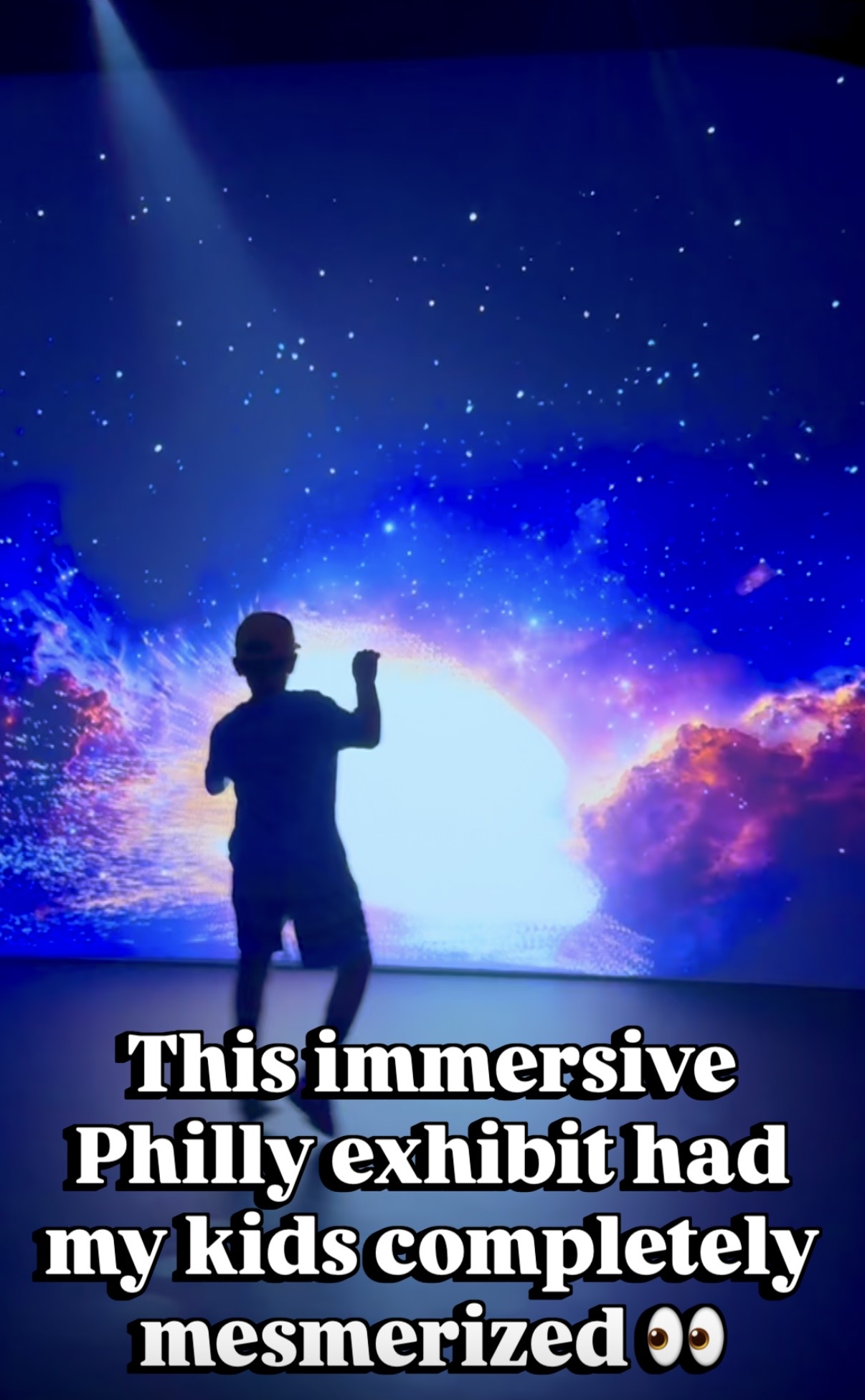 Child standing in front of a starry, colorful cosmic display with bright light, raising one hand. 20 words.