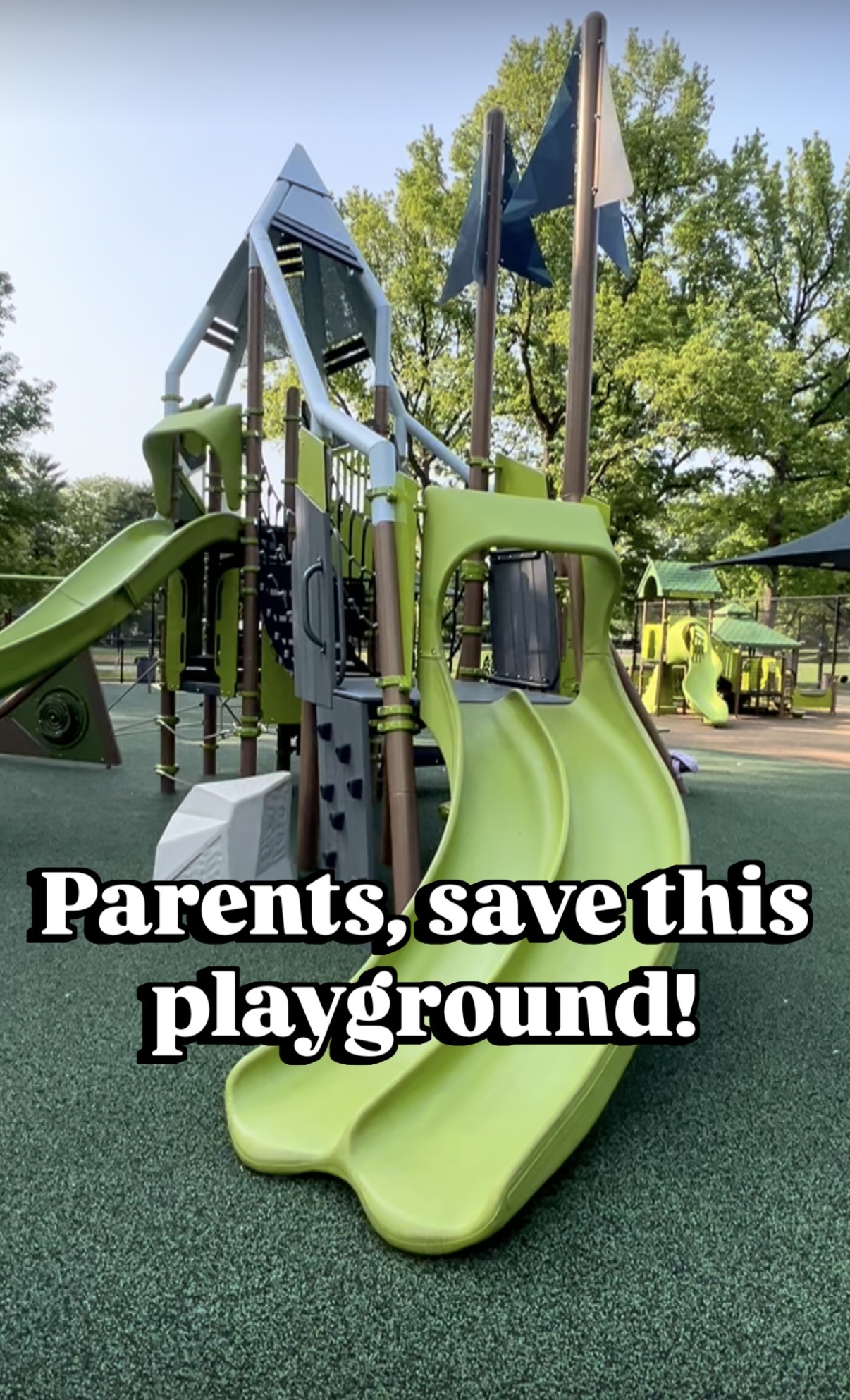 Children's playground with green slides, climbing structures, and flags, surrounded by trees and a clear sky.