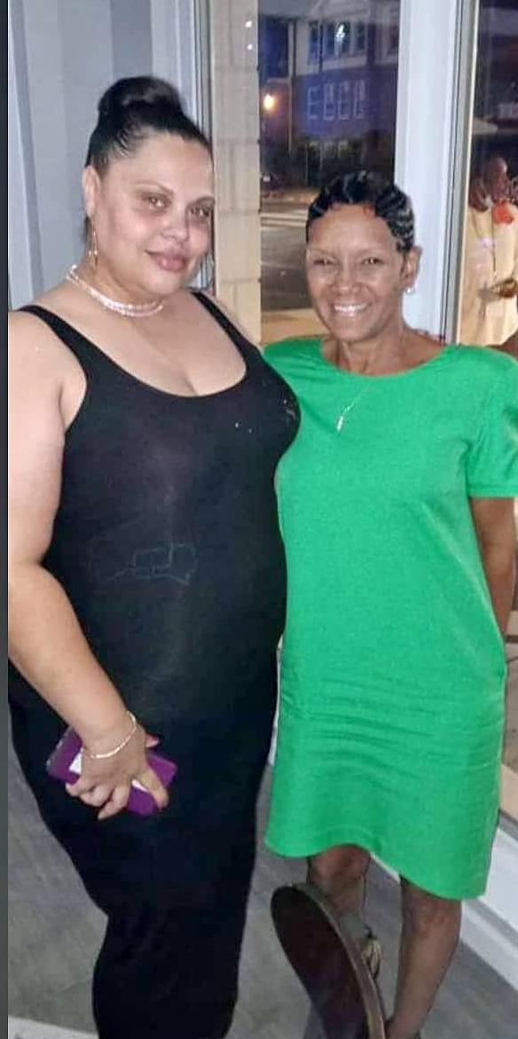 Two women standing indoors, one in a black dress and the other in a green dress, smiling at the camera.