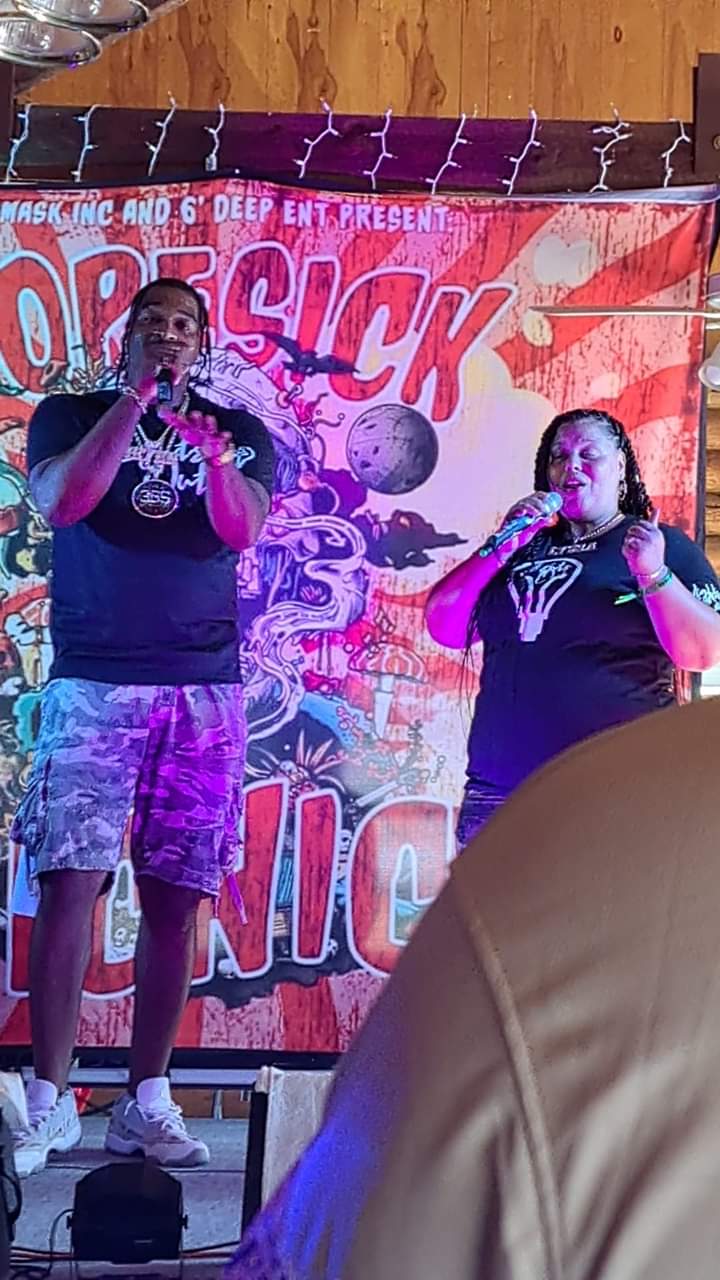 Two people speaking or singing on stage with a colorful backdrop, one holding a microphone, in a lively event setting.