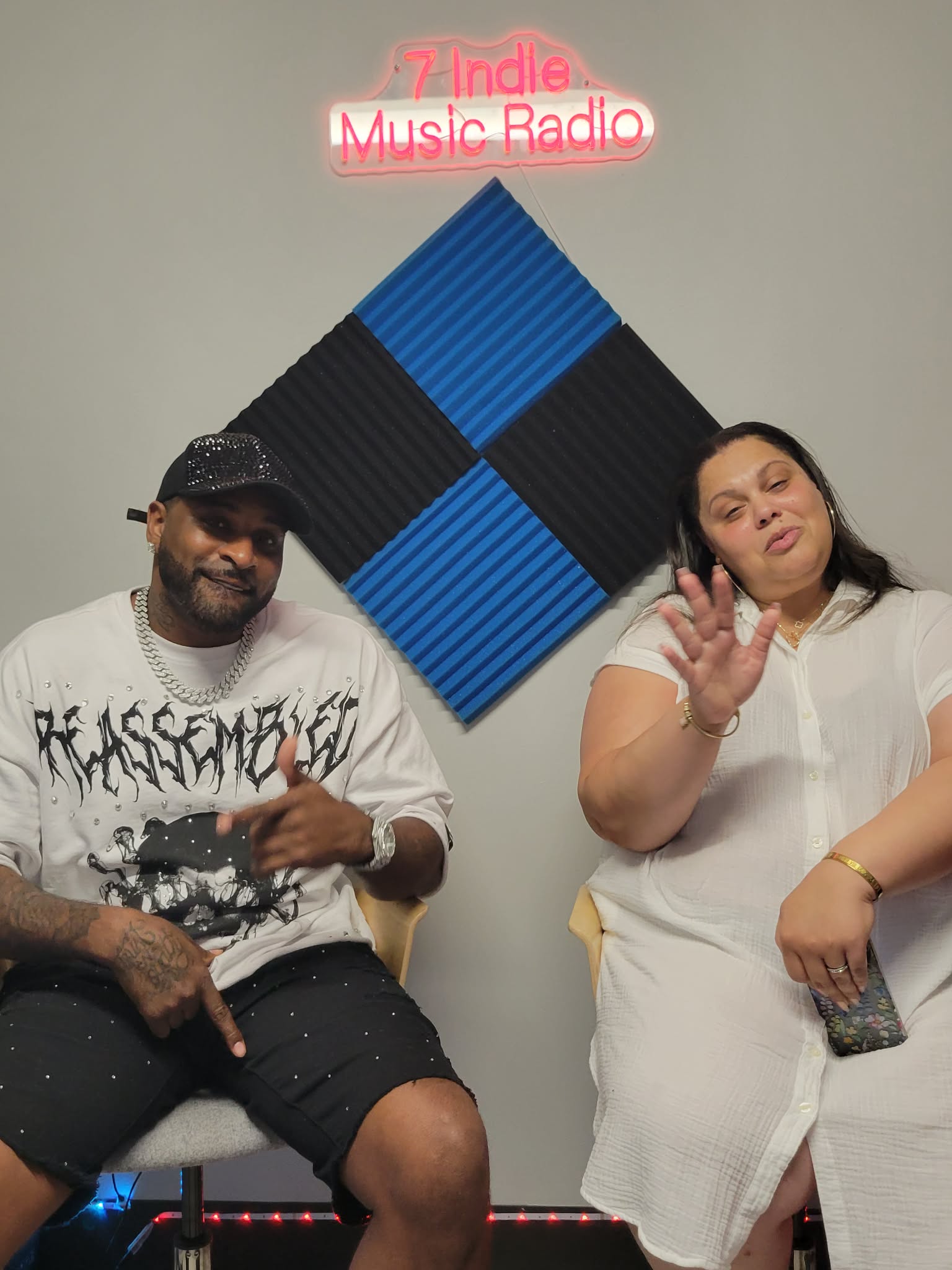 Two people sitting in front of a wall with a sign that reads '7 Indie Music Radio,' one making a peace sign and the other raising a hand.