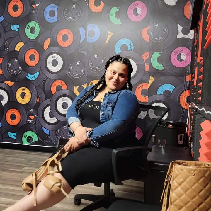 Woman sitting in a black chair with a colorful circular patterned wall behind her, holding a tan bag, smiling.