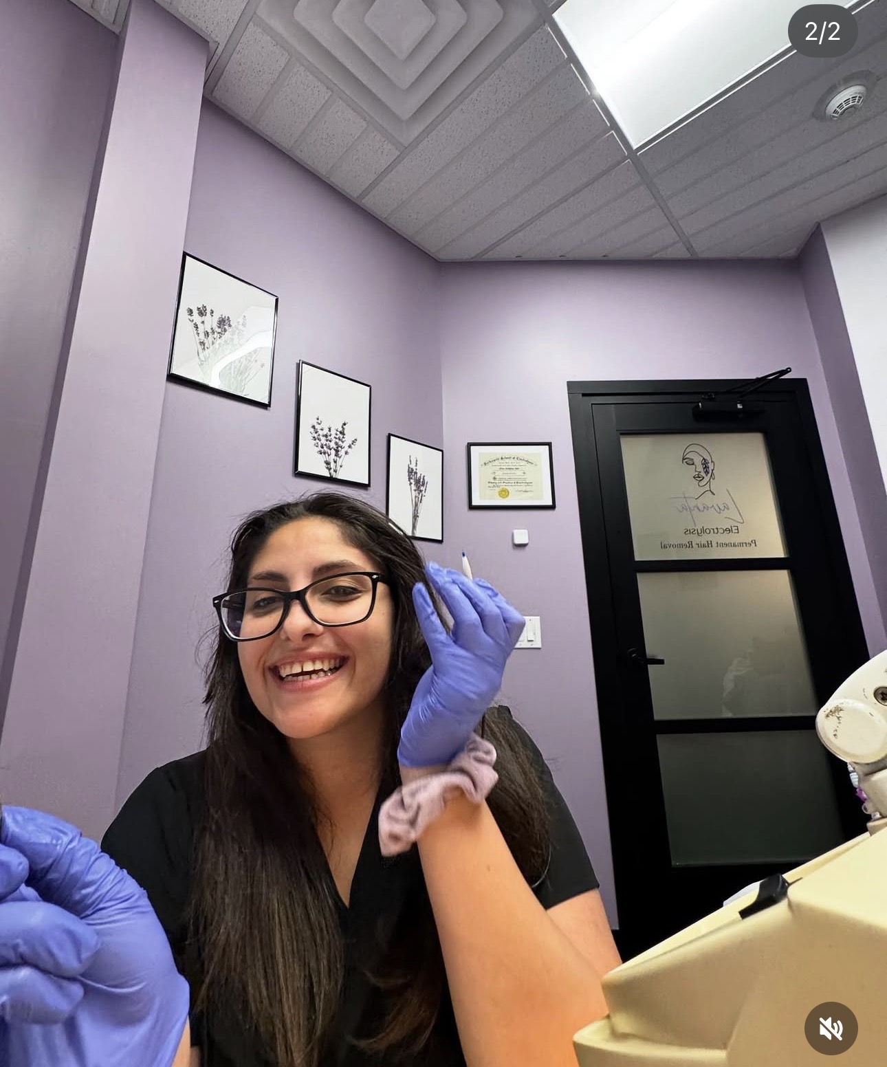 Smiling woman with glasses and long dark hair wearing purple gloves in a lavender-colored room with framed pictures on the wall.
