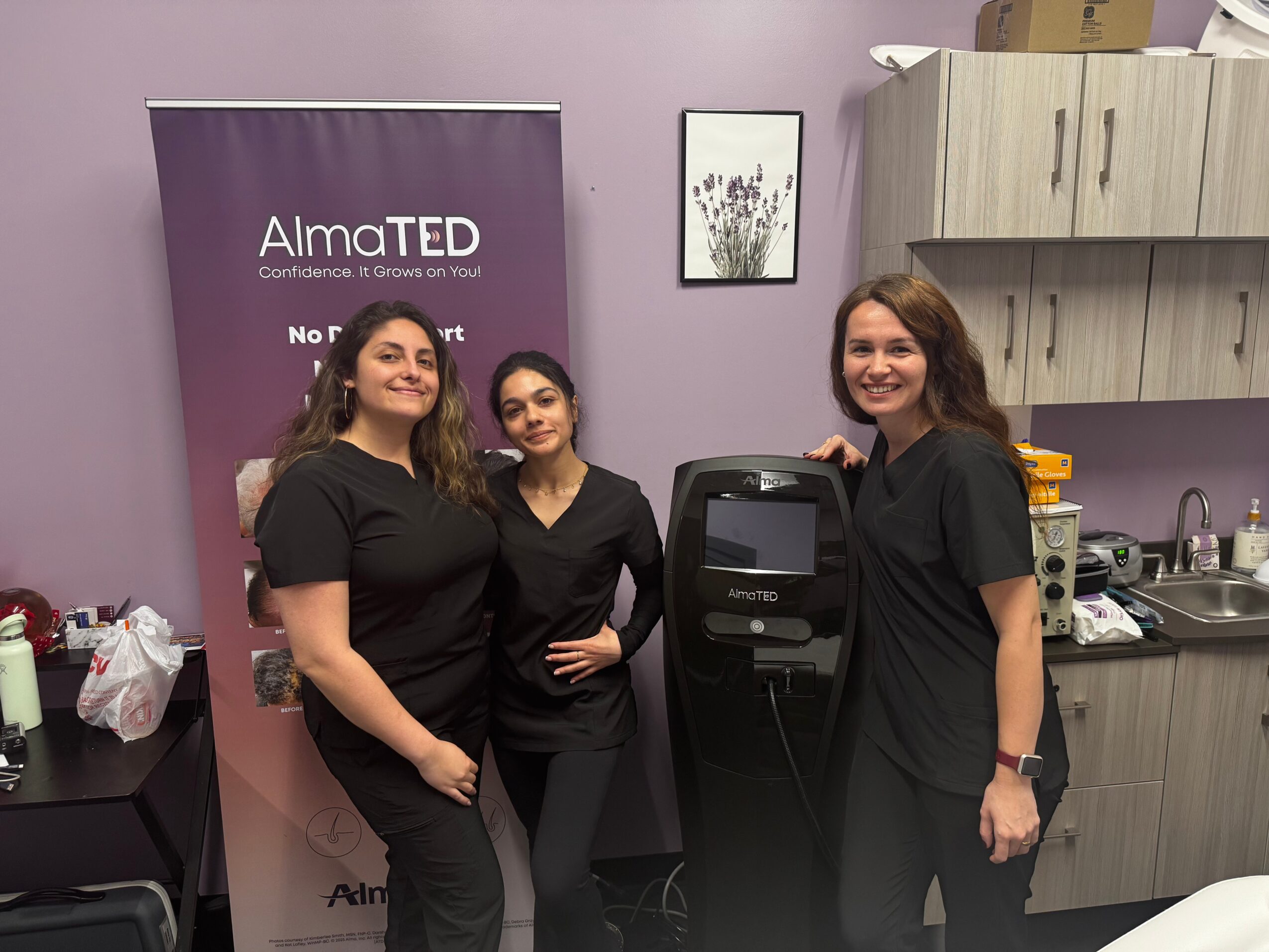 Three women in black uniforms standing in a room with a purple wall, a banner, and kitchen cabinets, smiling at the camera.
