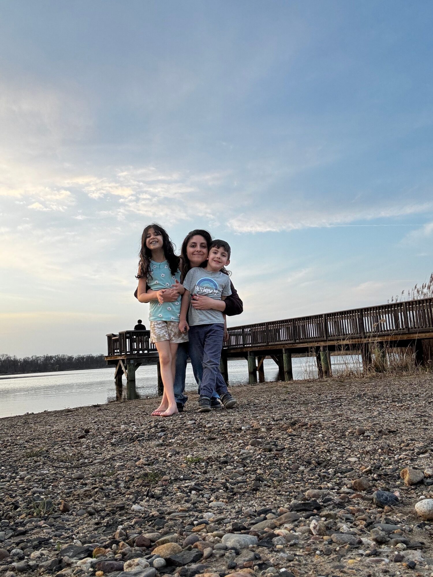 Three people standing on a rocky beach near a wooden pier, with water and sky in the background.