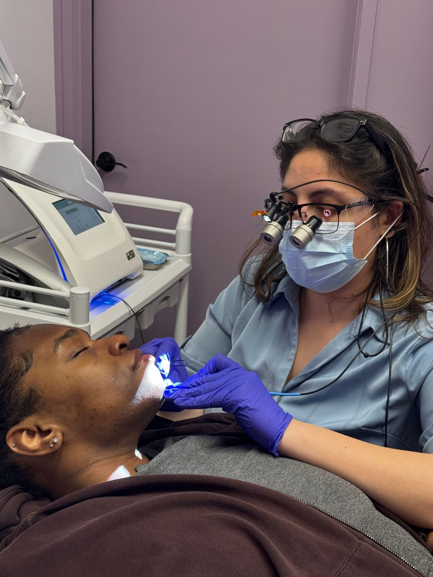 A healthcare professional wearing a mask, glasses, and gloves examines a patient lying on a bed.