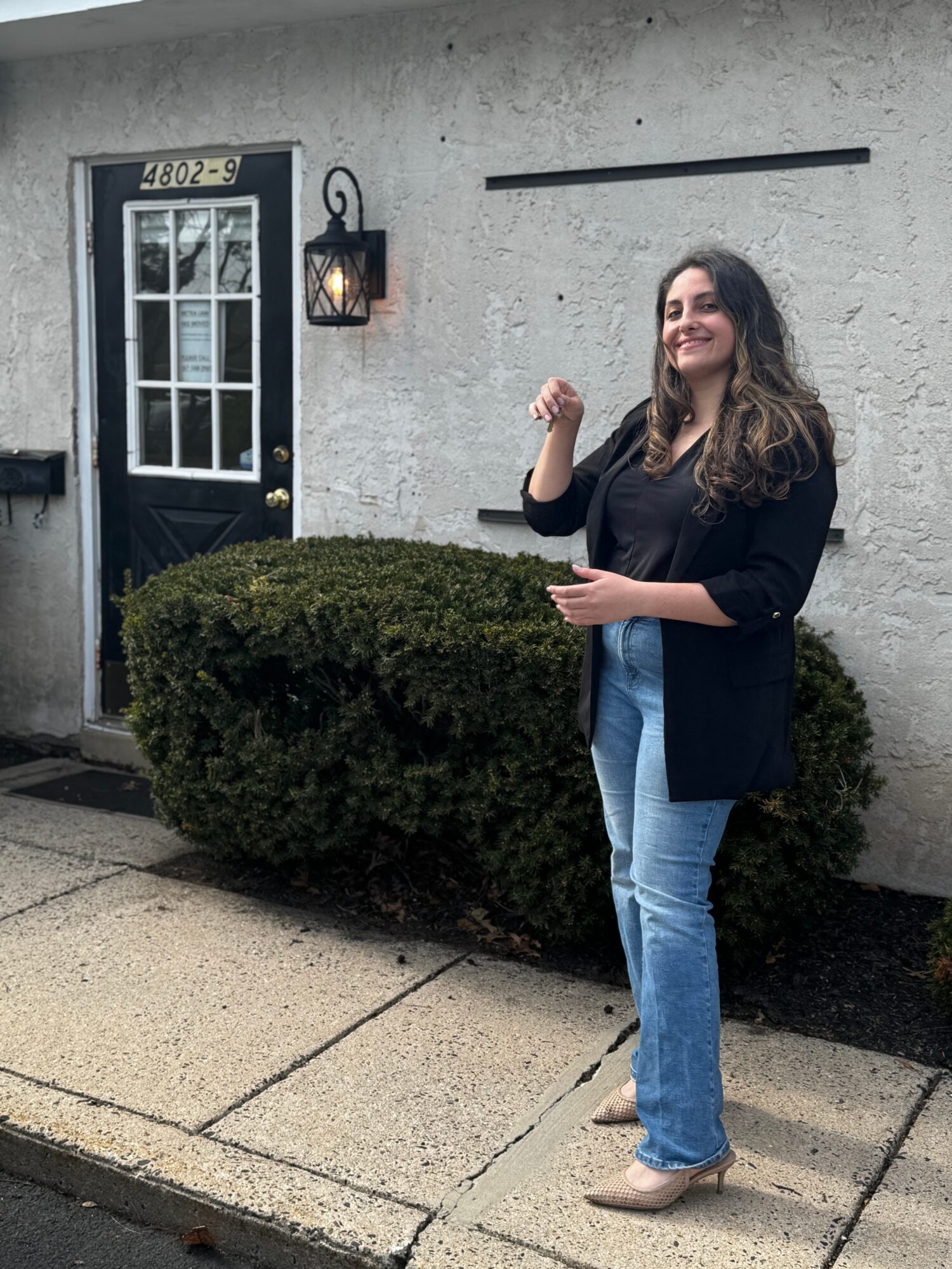 Woman with long hair smiling, wearing a black blazer and jeans, standing outside near a bush and a door.